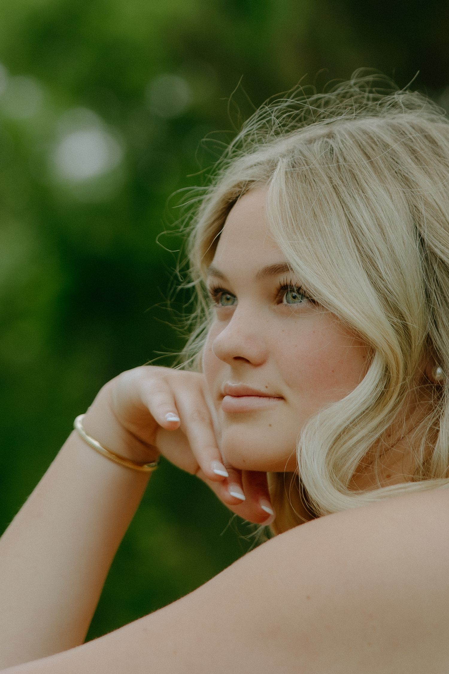 Young blonde woman with gold bracelet resting chin on hand, gazing softly with green bokeh background.