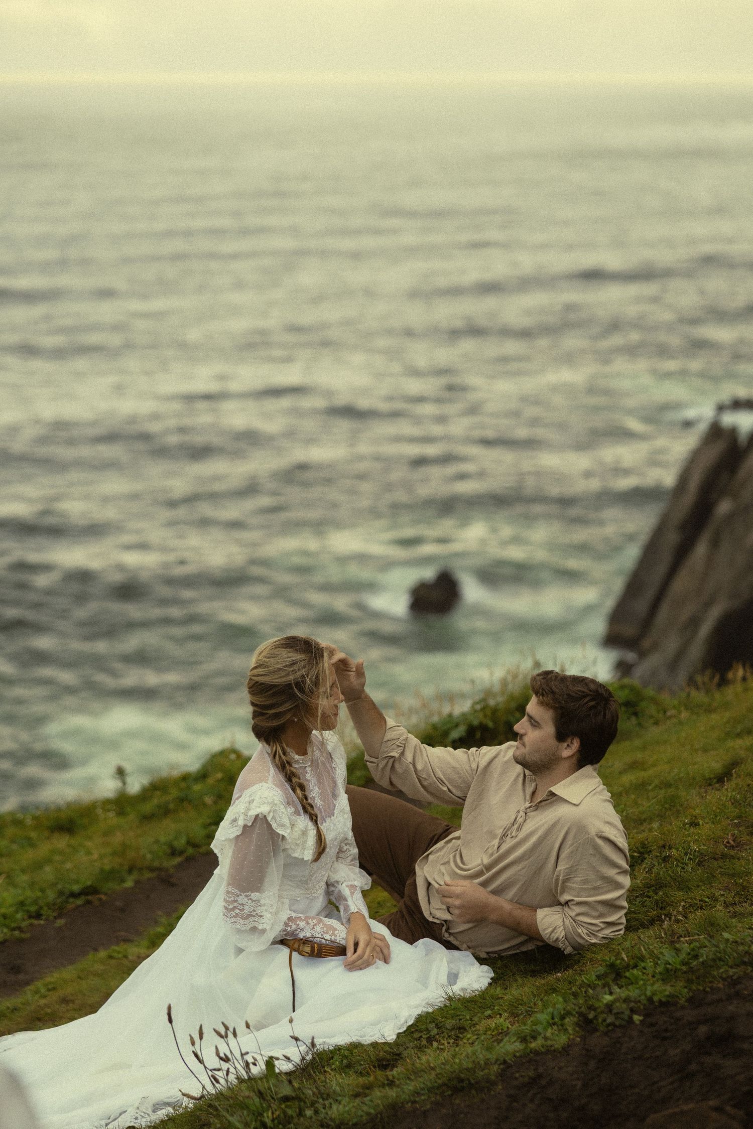 Couple sitting on coastal cliff, man gently touching woman's hair, ocean backdrop, romantic outdoor wedding photo.
