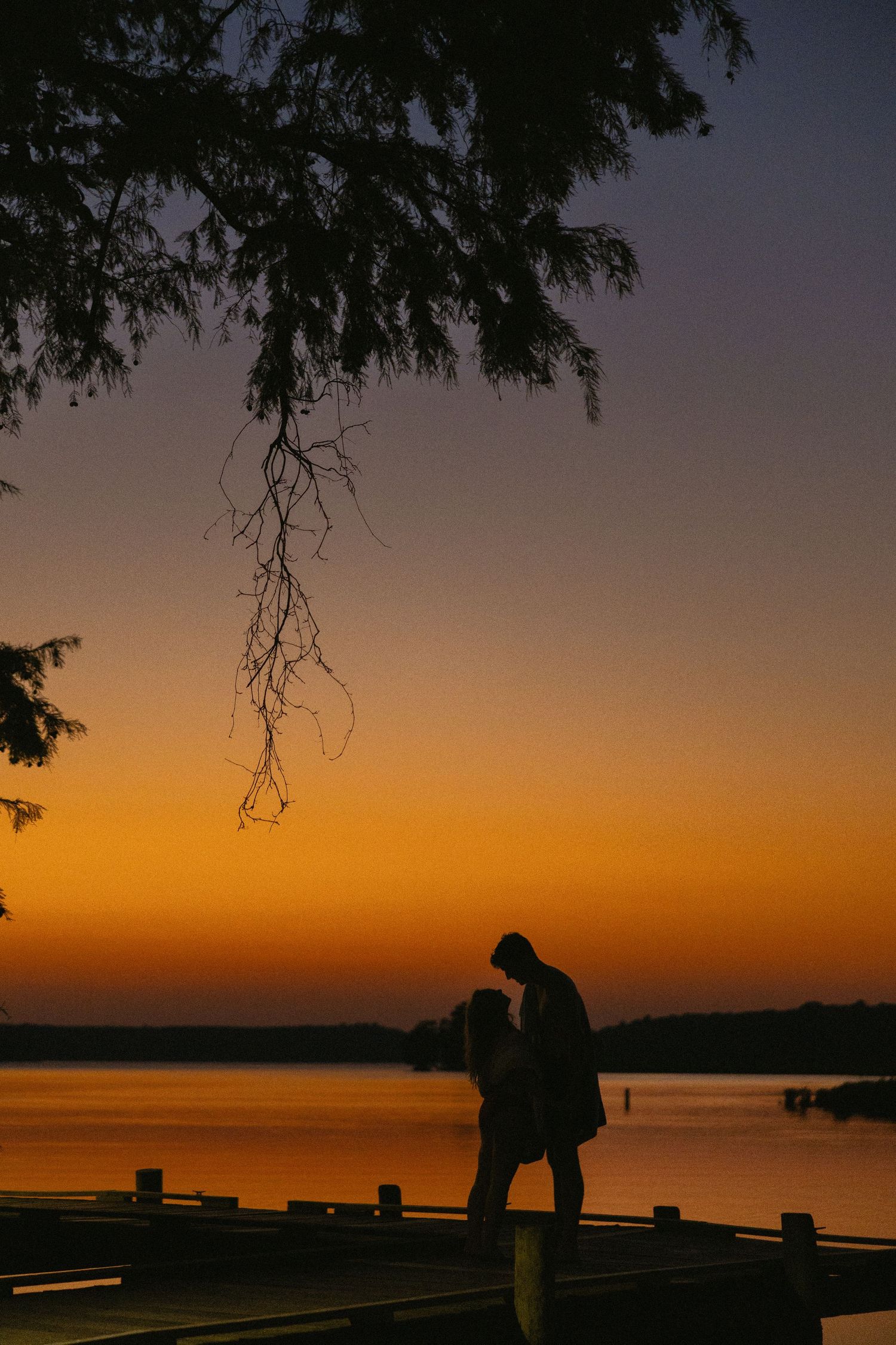 Romantic couple silhouetted against a vibrant orange and purple sunset on a lakeside dock with tree branches overhead.