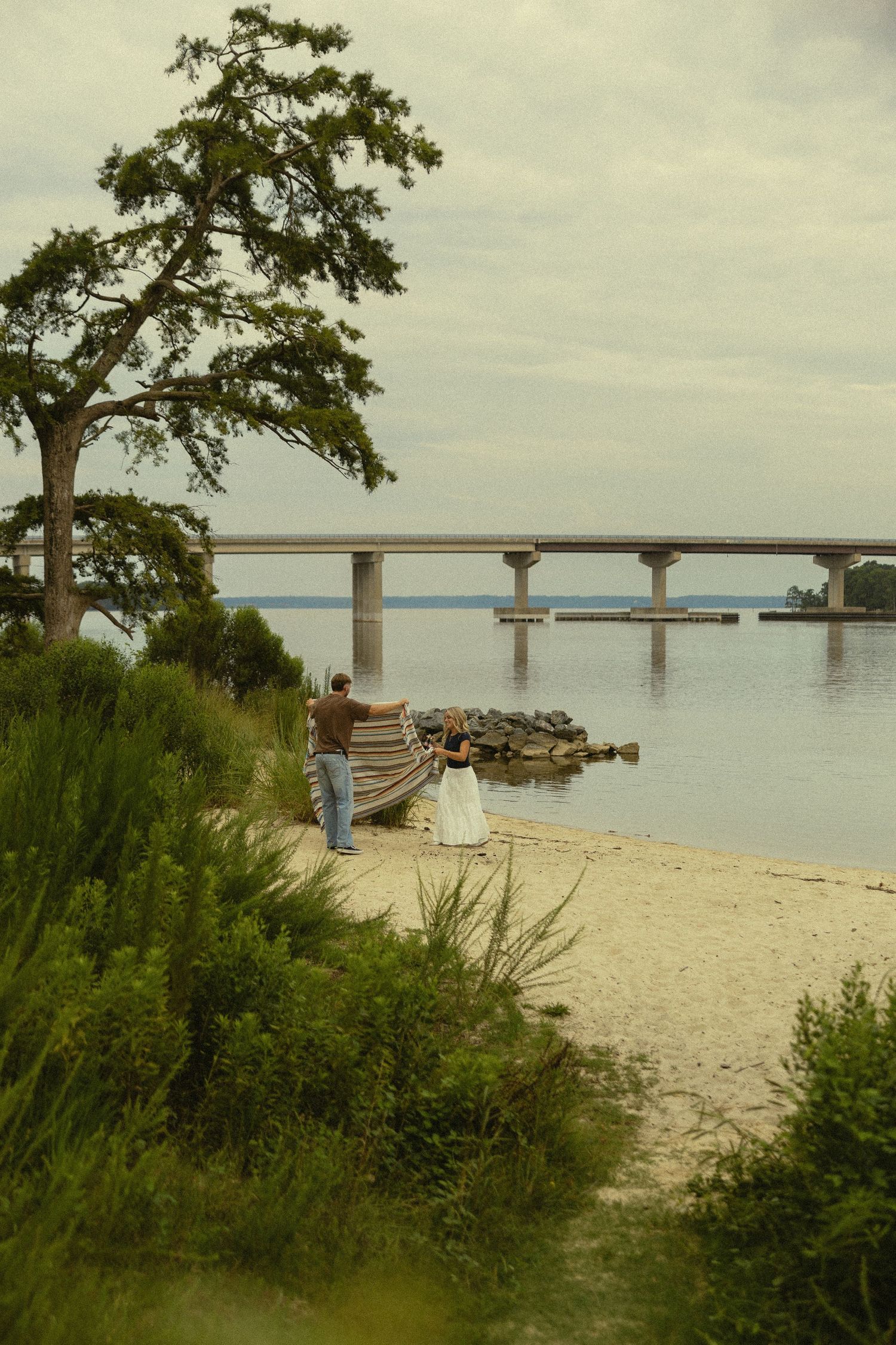 Couple in wedding attire standing on sandy beach by calm water with a large bridge and green tree in background.