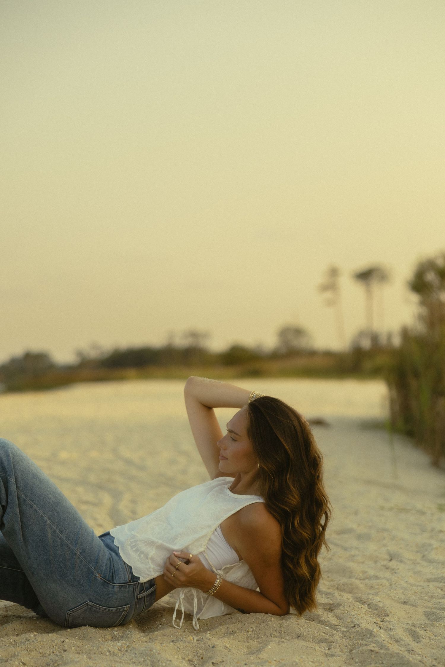 Woman in white top and jeans relaxing on sandy beach at sunset with palm trees in background.