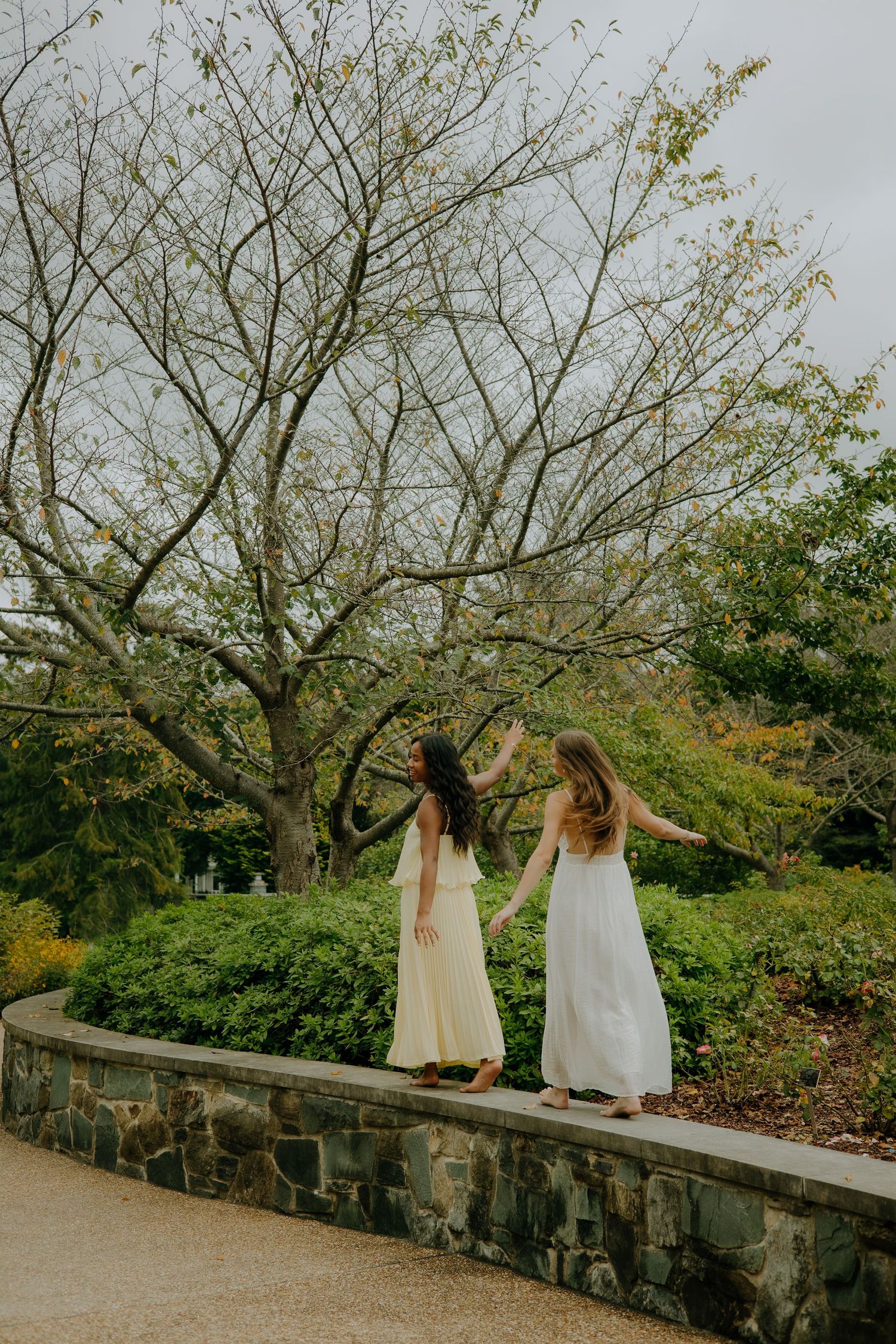 Two people in light dresses stand on a stone wall surrounded by spring trees.