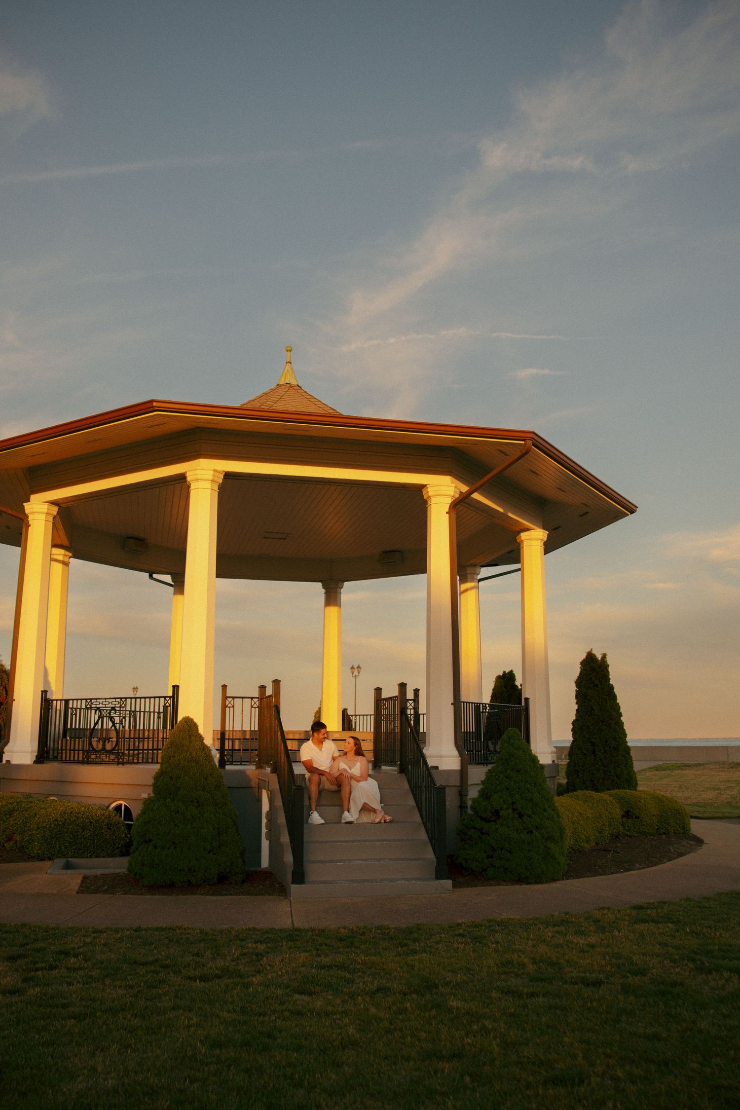 Couple sitting on steps of illuminated gazebo at sunset with manicured shrubs and ocean view in background.