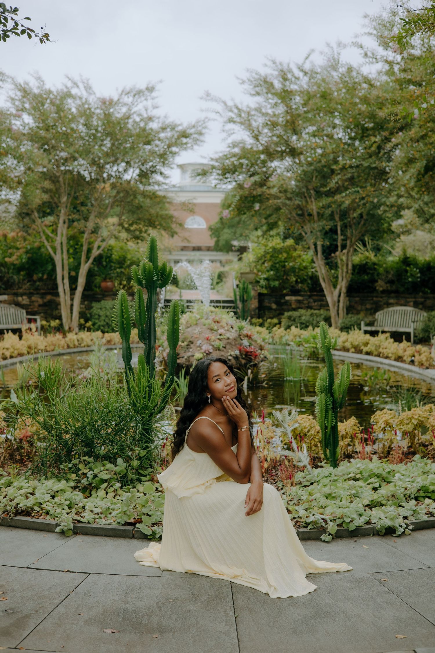 A woman in a yellow dress sits gracefully in a lush botanical garden with cacti and a pond.