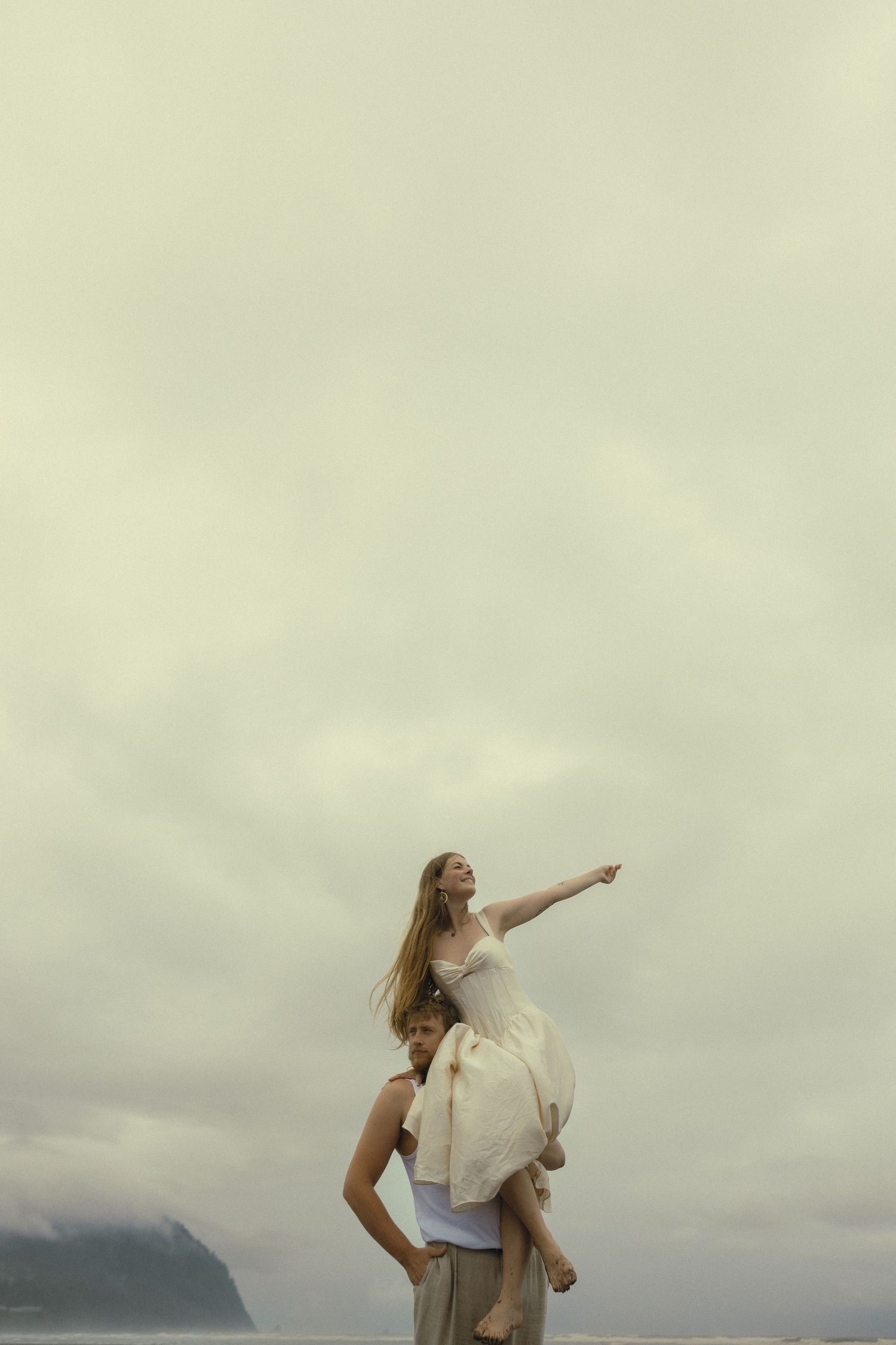 Woman sitting on man's shoulders pointing at cloudy sky near coastal cliffs in romantic outdoor portrait.