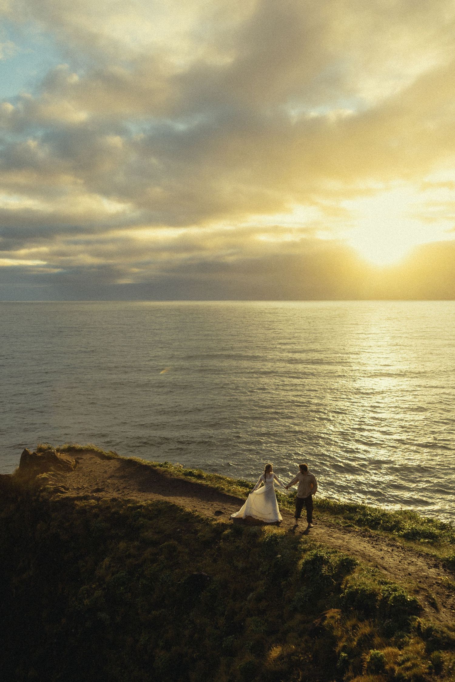 Couple in wedding attire holding hands on a coastal cliff at golden sunset with dramatic clouds over the ocean.