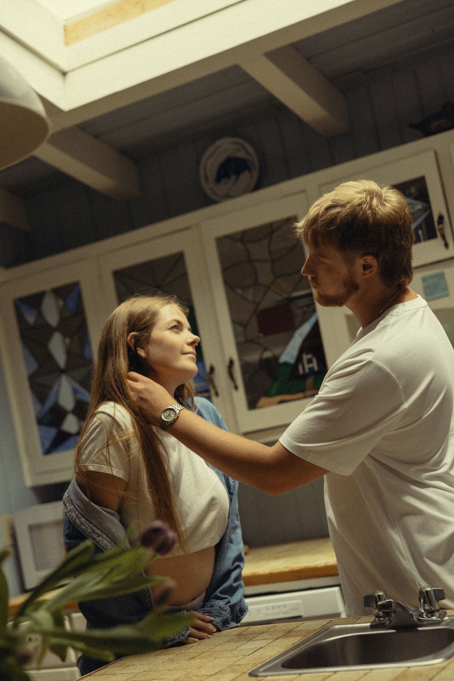 A young couple shares a tender moment in a rustic kitchen, with the man gently touching the woman's hair.