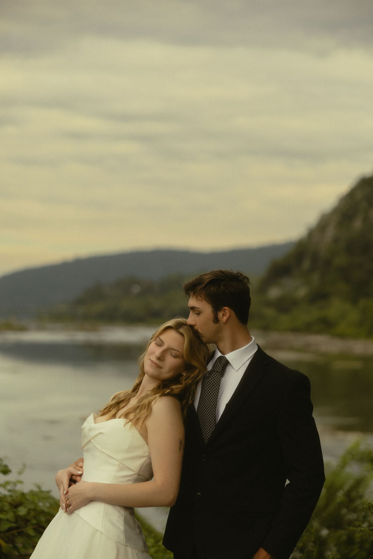 Bride and groom embrace outdoors near a lake with misty mountains, wearing wedding attire in a romantic portrait.