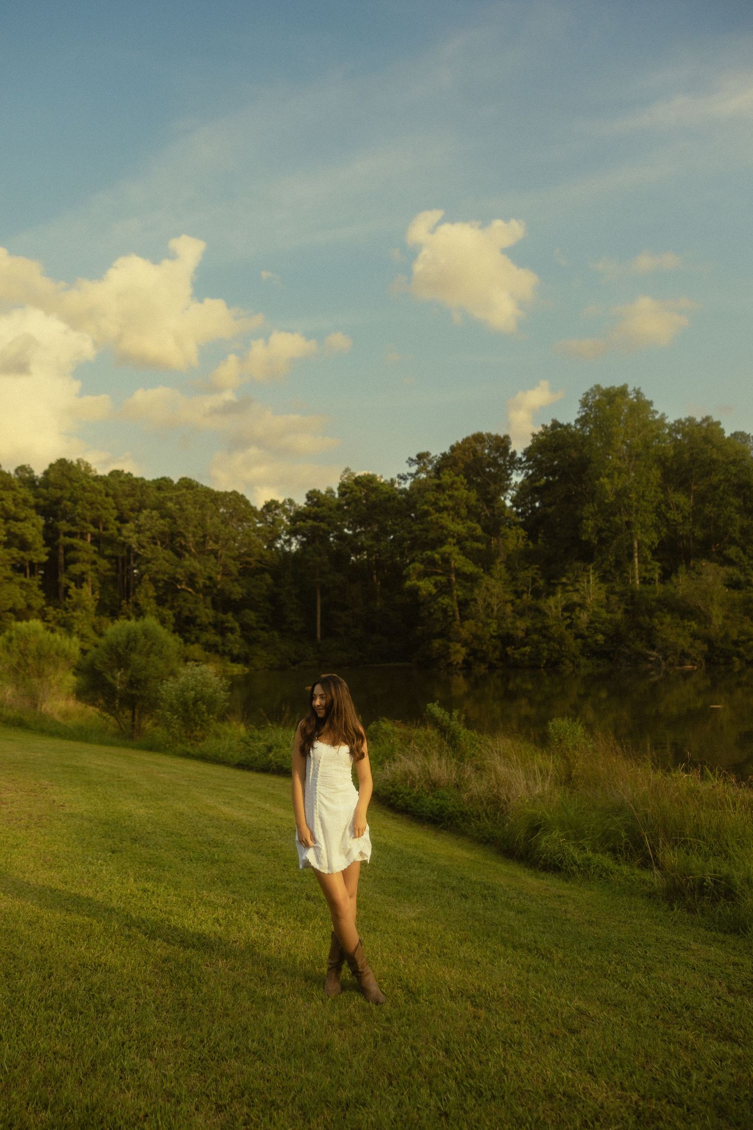 Young woman in white dress and cowboy boots standing on green grass near a pond with trees and blue sky.