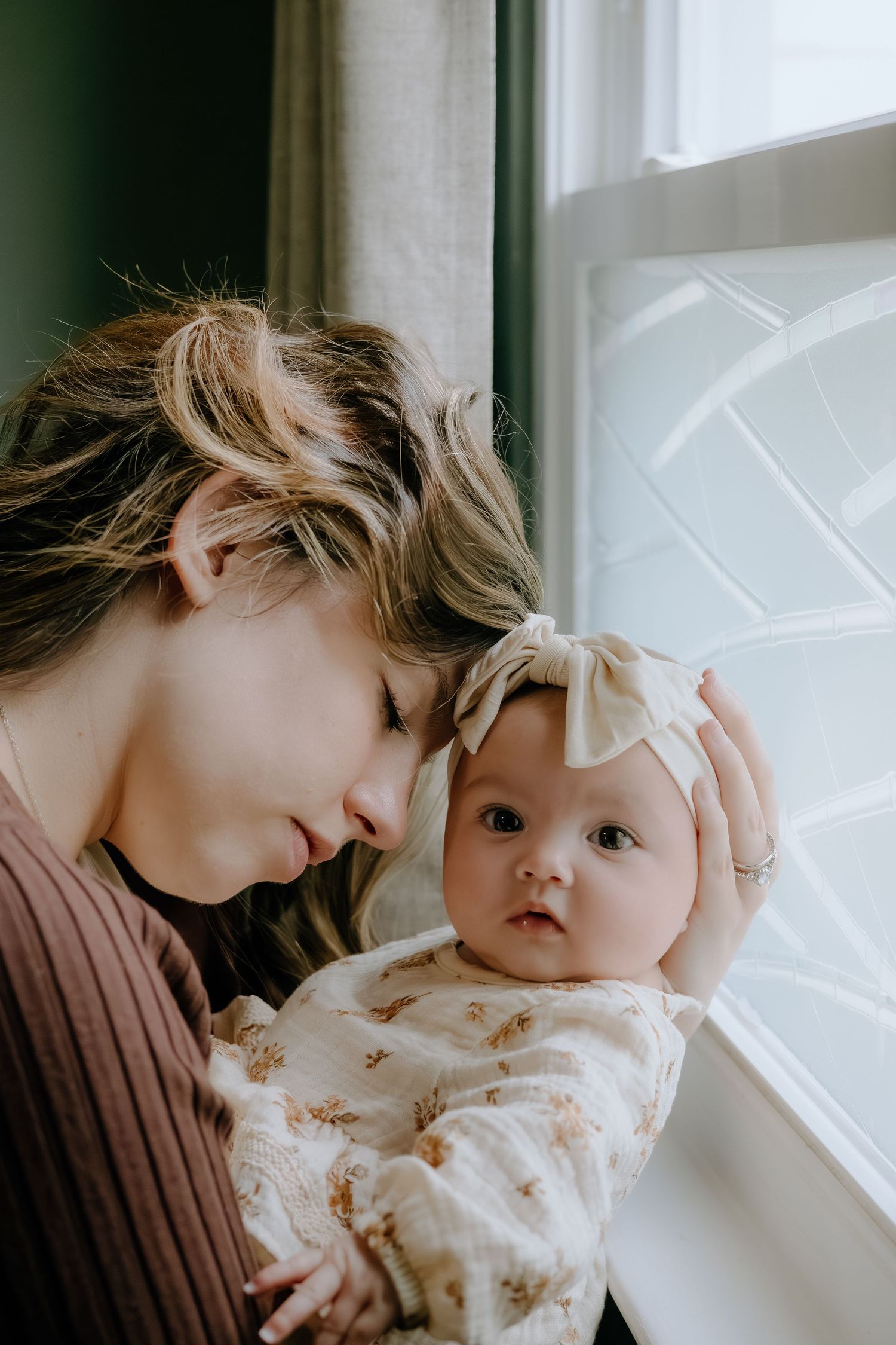 Tender moment between baby in floral dress and caregiver near a bright window.