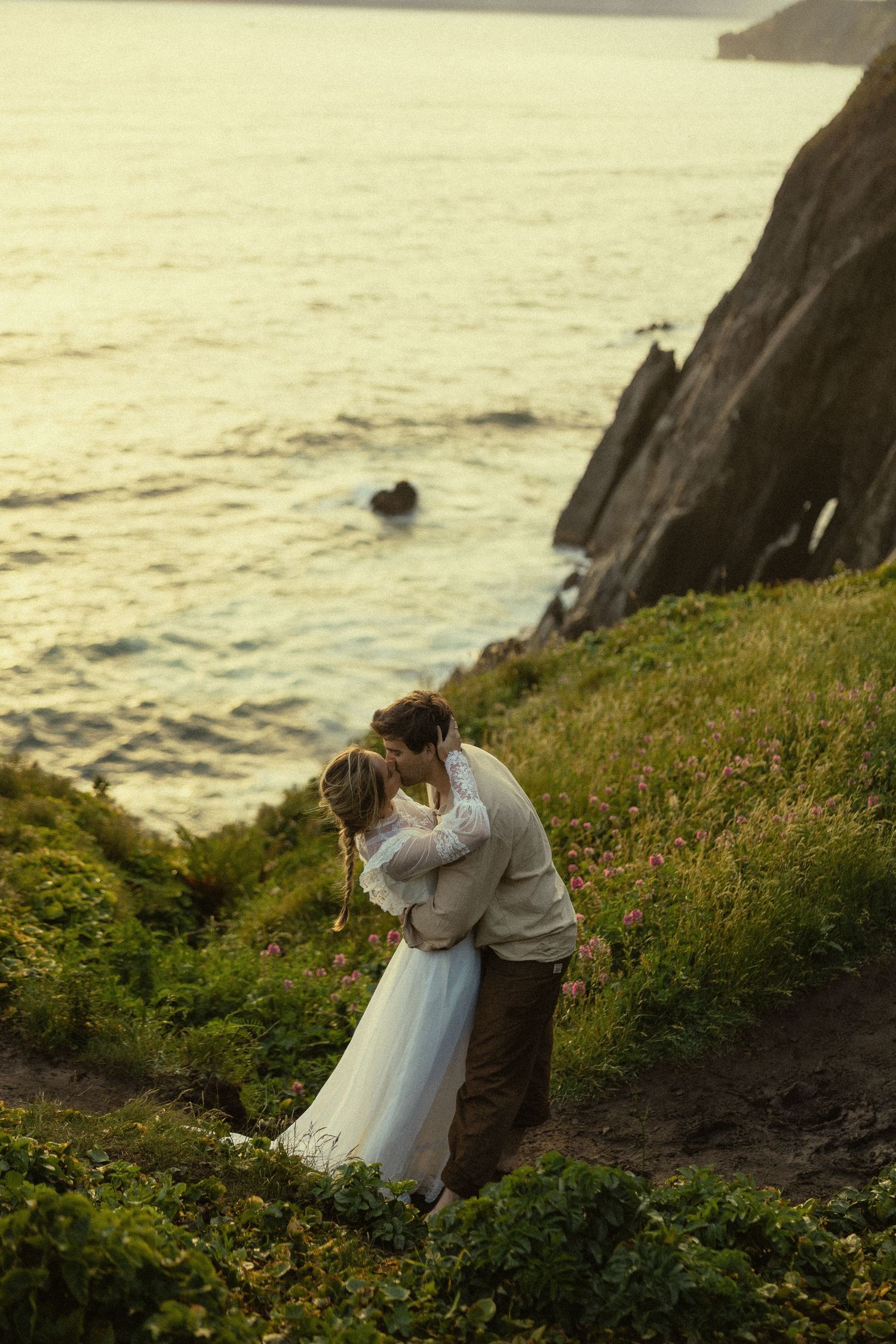Couple embracing on a cliffside overlooking the ocean at sunset, bride in white dress and groom in tan suit.