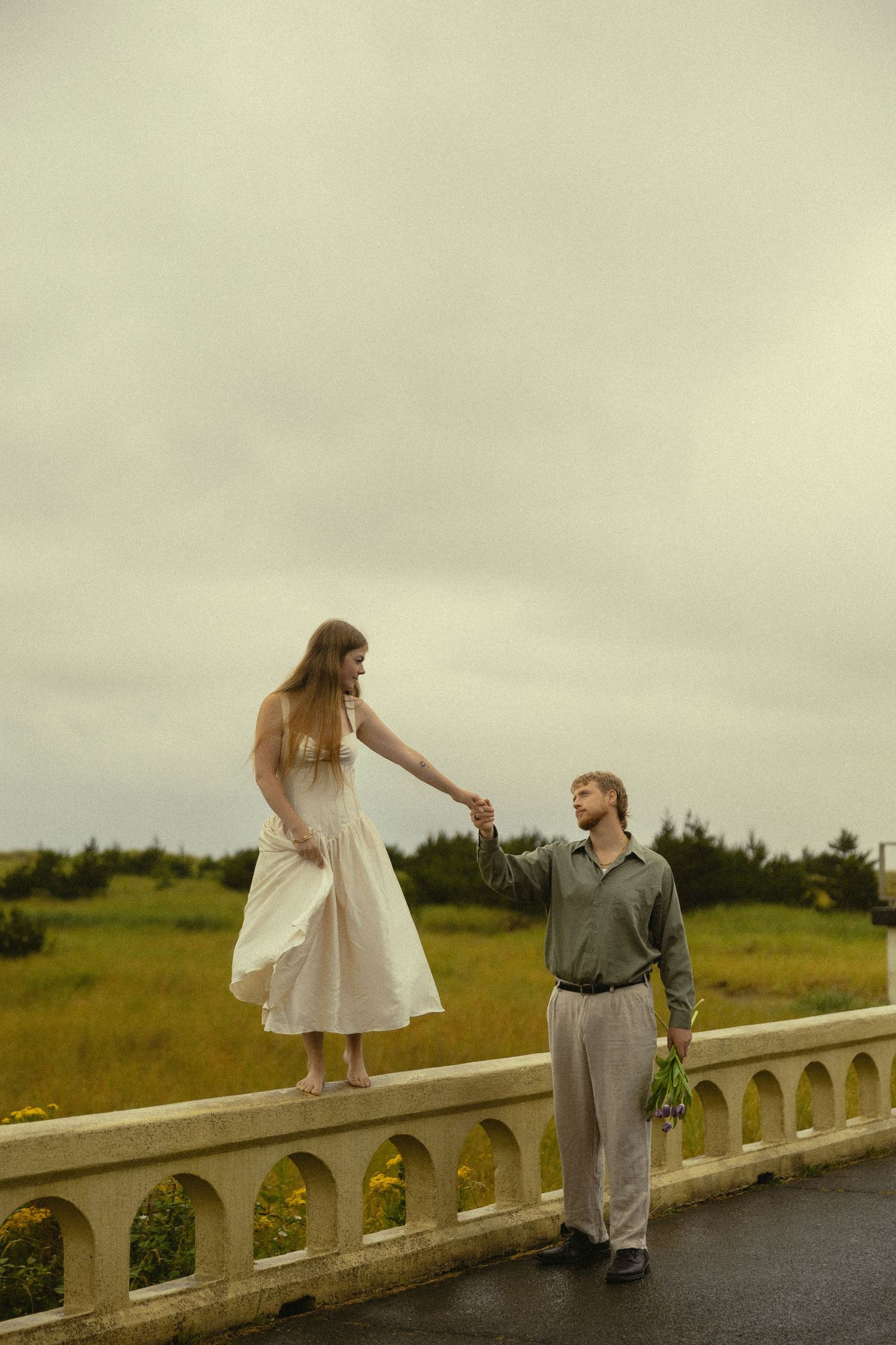 A woman in a white dress stands on a bridge railing holding hands with a man in a field.