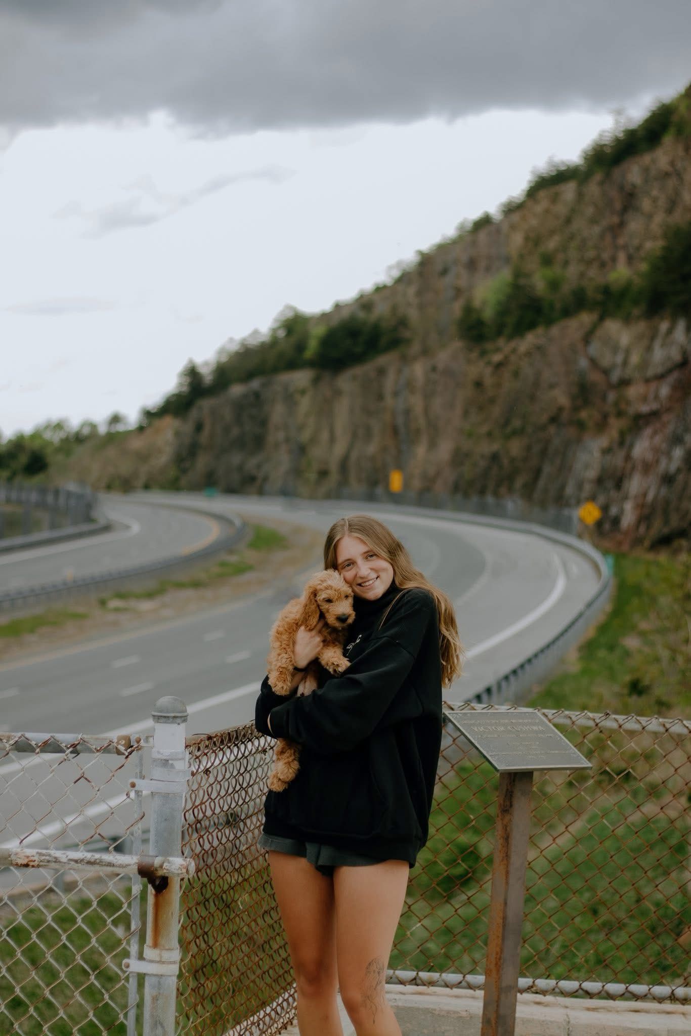 Young woman in black hoodie and shorts smiling while holding a golden puppy near a winding mountain road.
