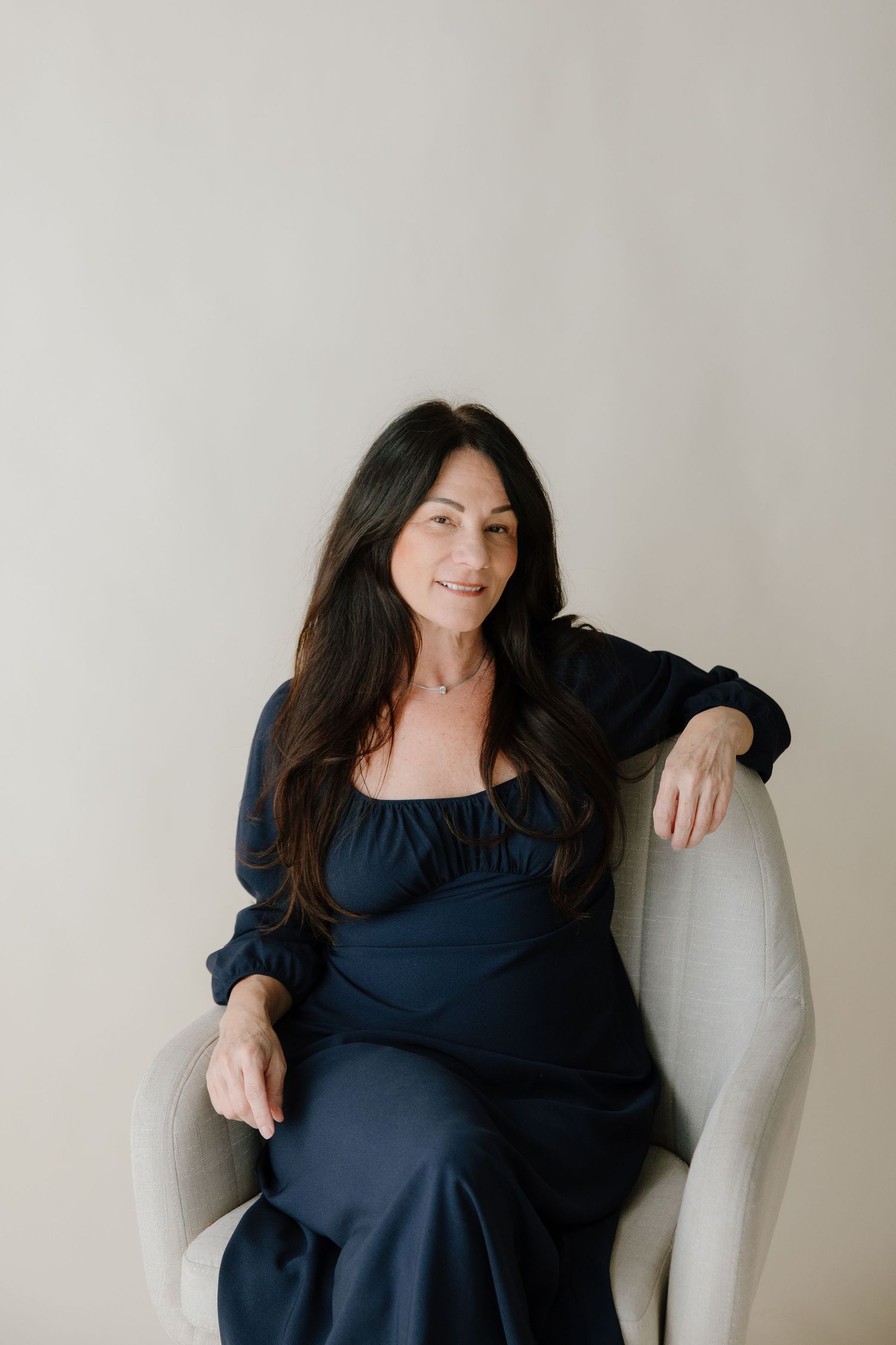 Woman in navy blue dress lounges comfortably on light gray armchair in minimalist setting.