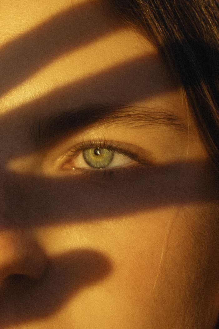 Close-up of a green eye with dramatic shadow stripes across a woman's face in warm golden light.