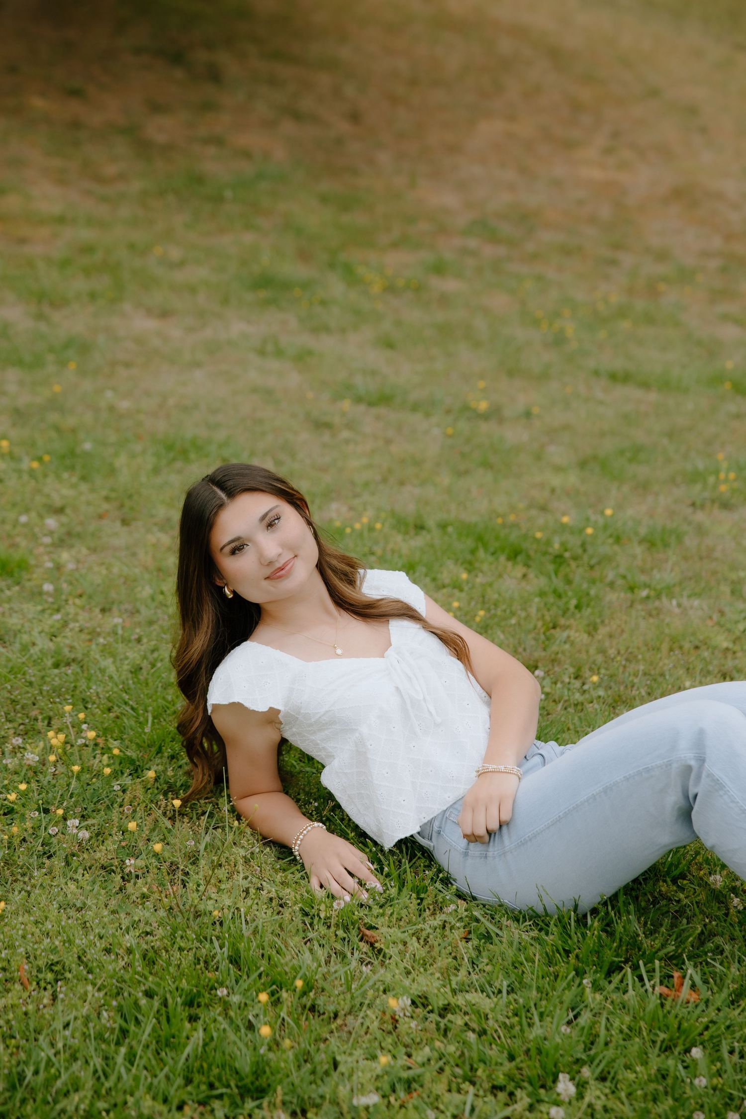 Young woman in white top and jeans relaxing on green grass, smiling softly in a sunny outdoor setting.