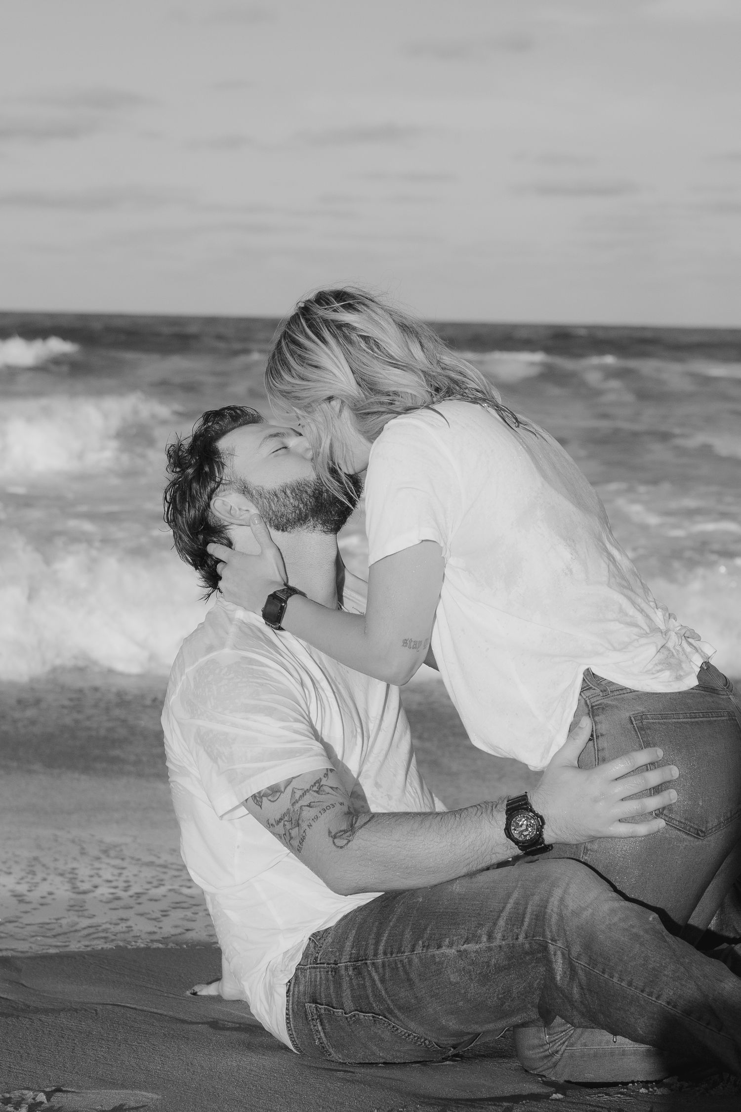 Black and white photo of a romantic couple kissing on a beach with ocean waves in the background.