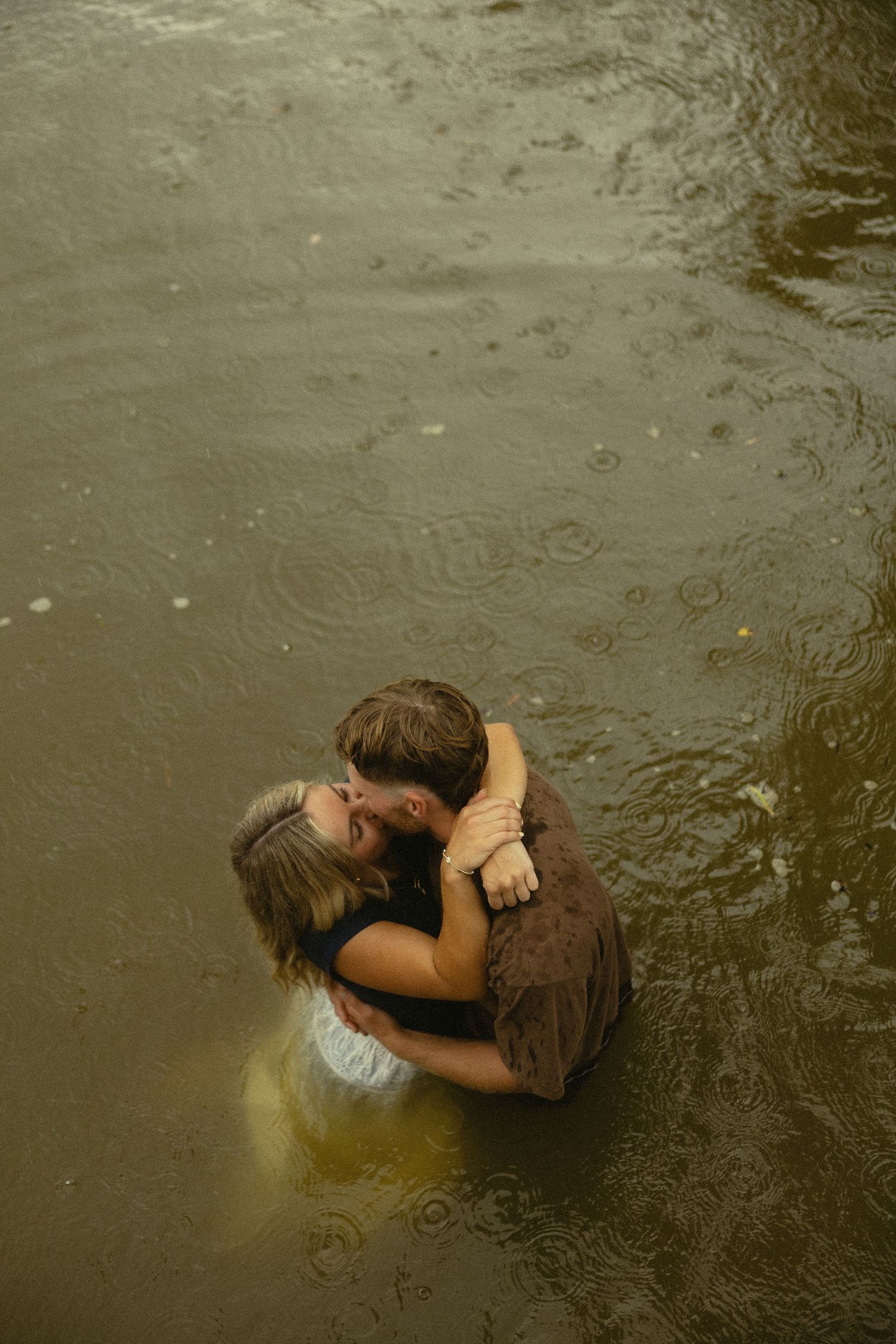 Couple sharing a romantic kiss while standing in a shallow river during rainfall.