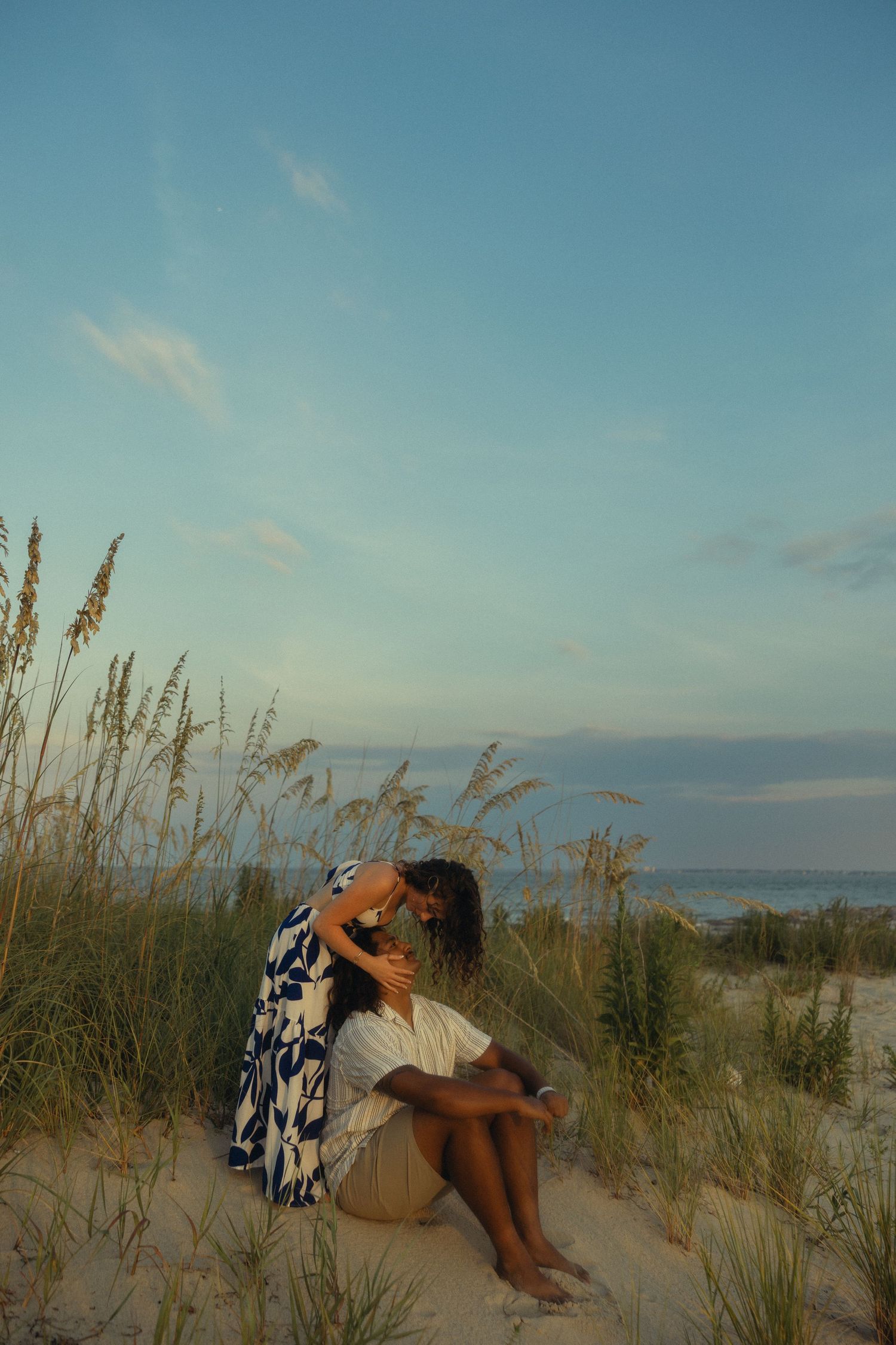 Two people embrace on sandy beach dunes at sunset, surrounded by sea oats with ocean in background.