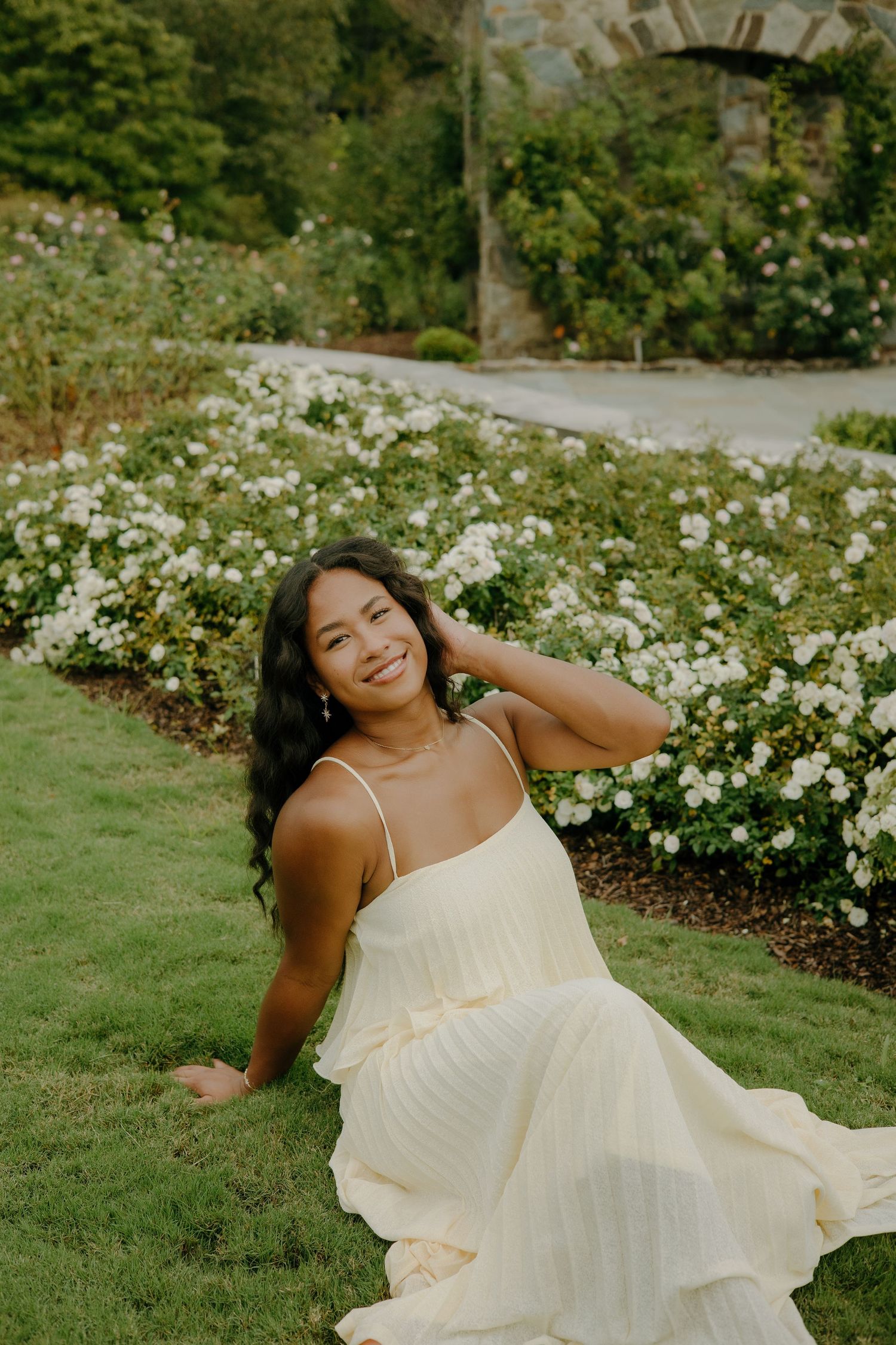 Person in flowing white dress posing gracefully on grass near white flowers.