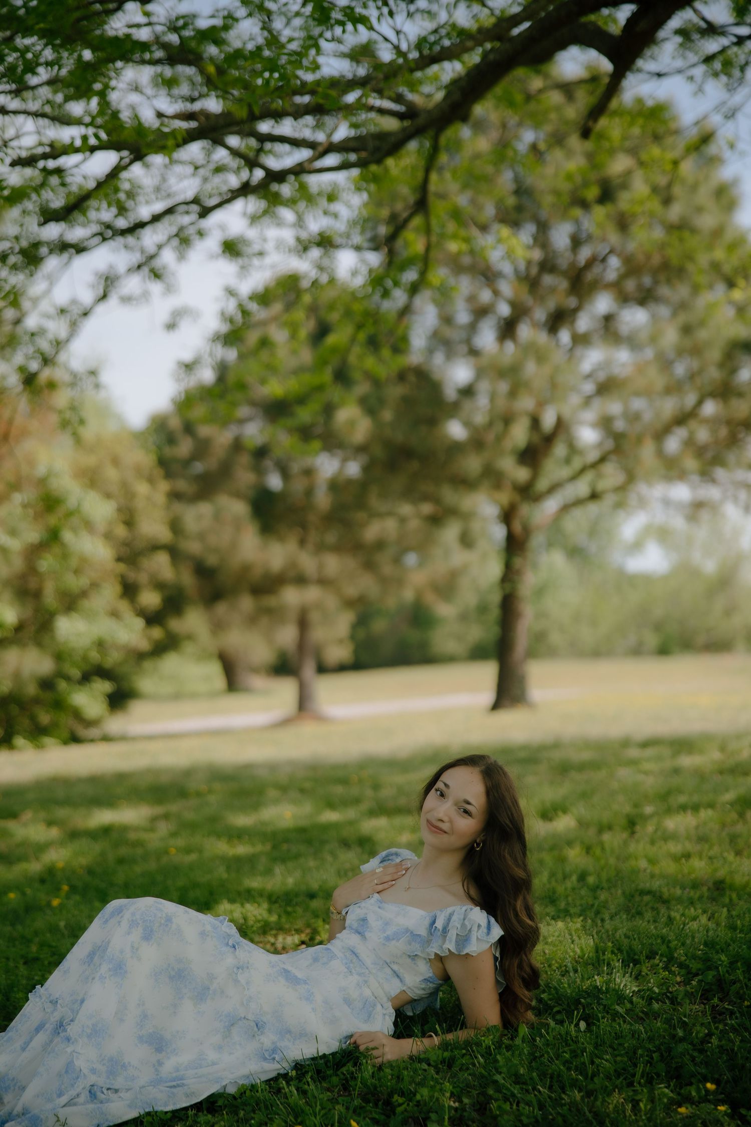 Pregnant woman in blue floral dress reclining on green grass under trees in a serene park setting.