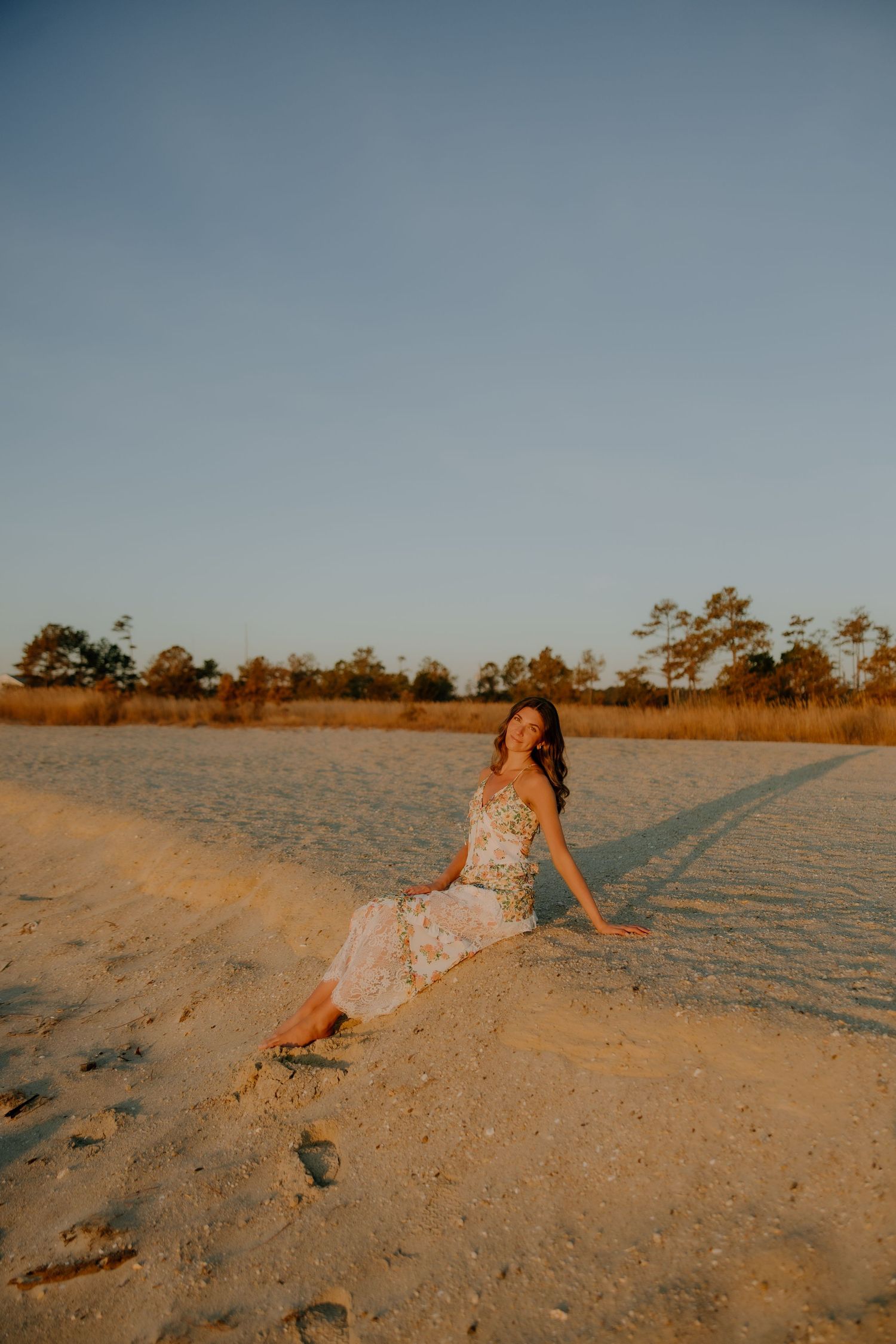 Woman in floral dress sitting on sandy ground at golden hour with trees in background.