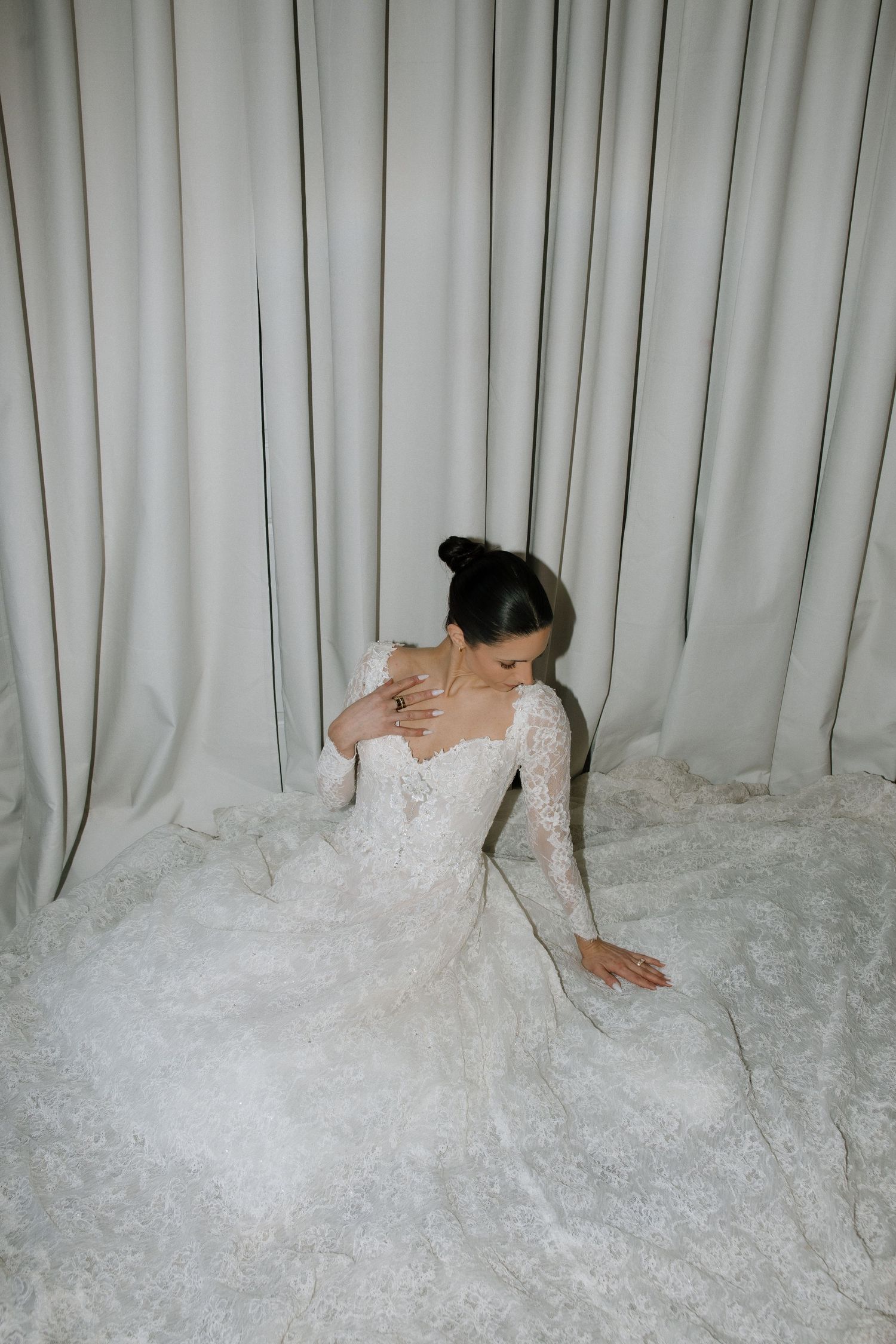 Bride in long-sleeve lace wedding gown with cathedral train sits gracefully on floor against soft white draped curtains.