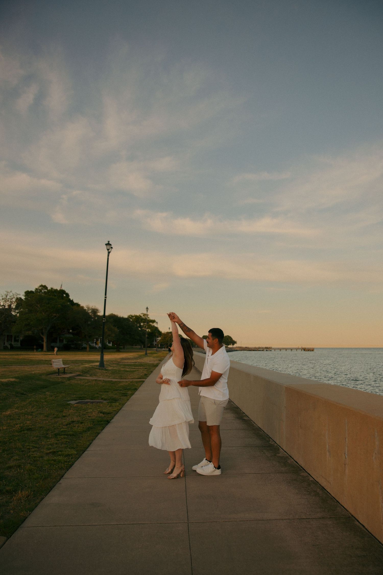 Couple in white outfits dancing on a waterfront path at sunset, with a park and pier in the background.