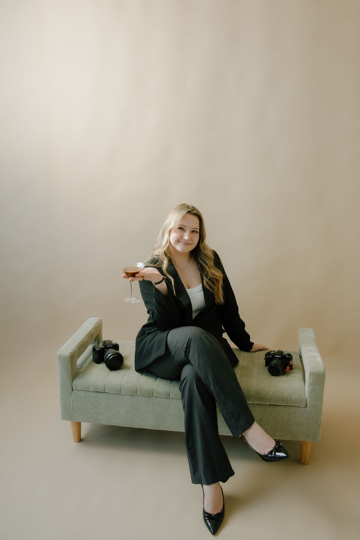 Blonde woman in black suit holding wine glass seated on gray bench with cameras, studio portrait photography.