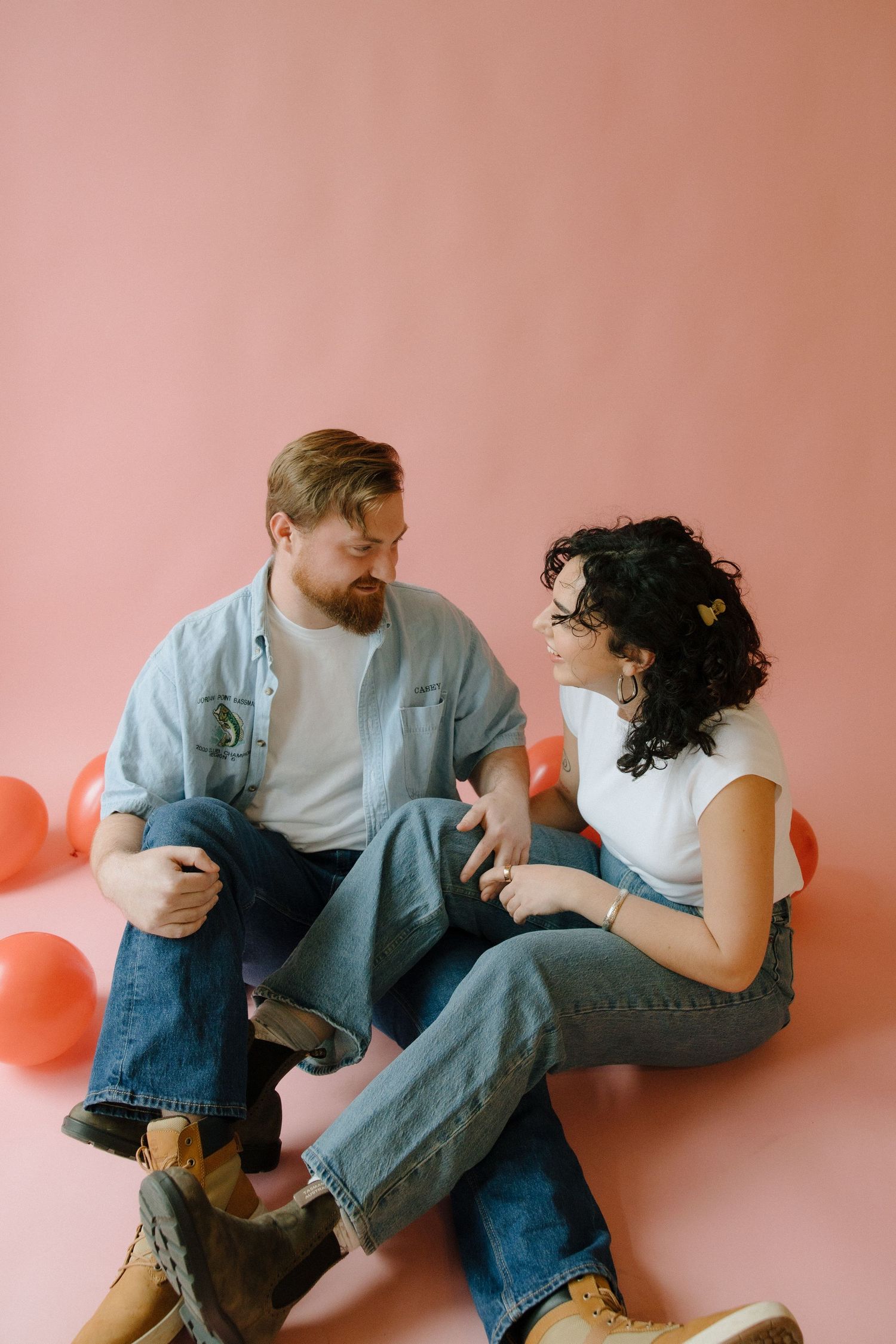 Couple sitting on pink floor with red balloons, wearing jeans and casual tops, smiling and talking to each other.