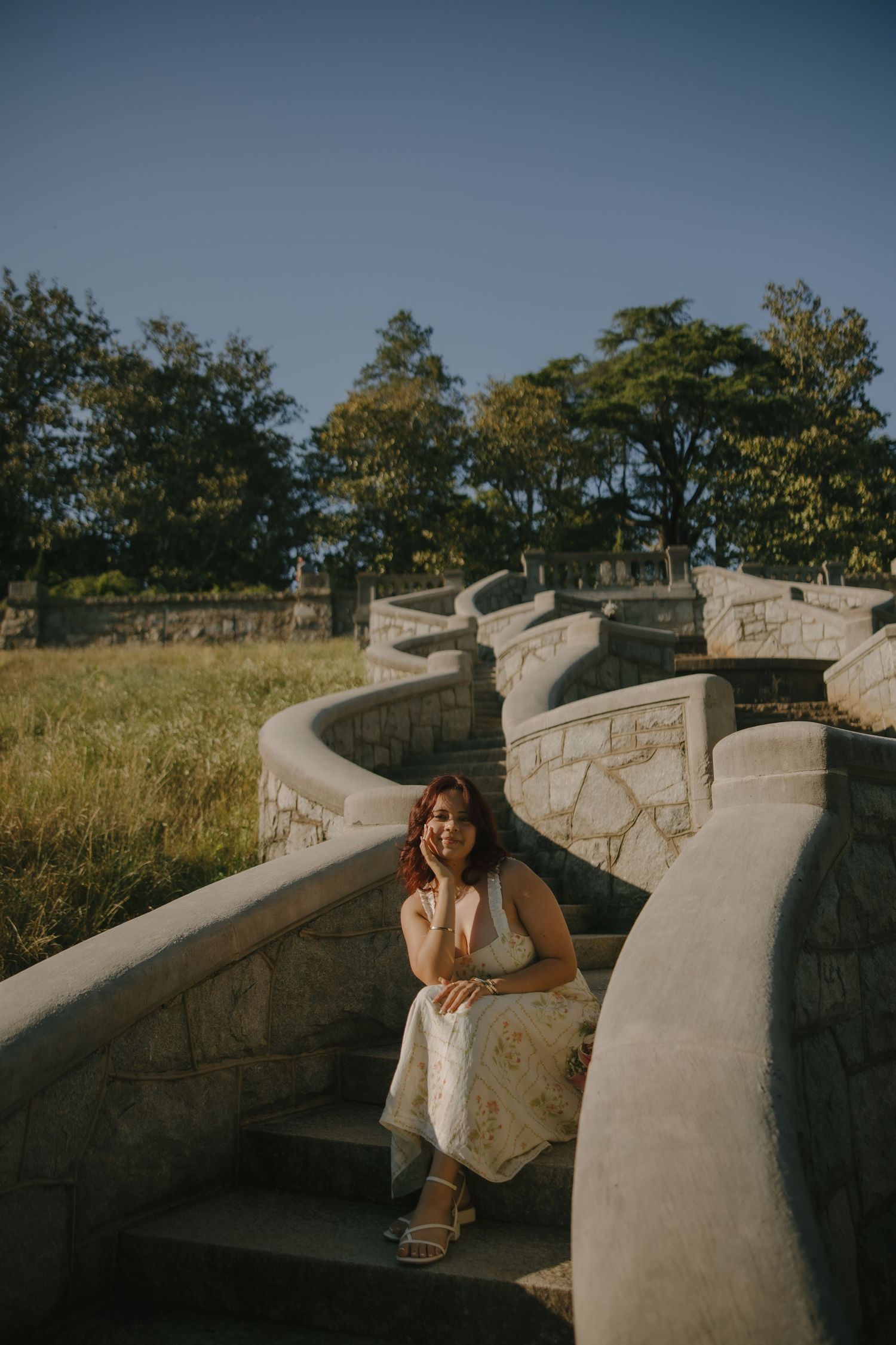 Woman in floral dress sitting on stone steps outdoors, surrounded by green trees and golden grass under a clear blue sky.