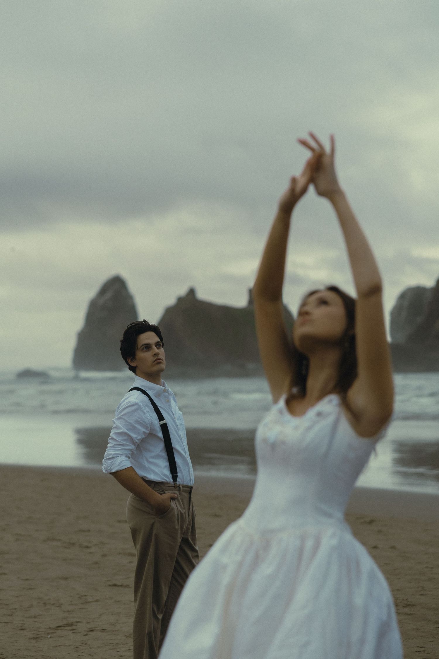 Couple on a moody beach, woman in white dress with arms raised, man in suspenders gazing at rocky ocean backdrop.