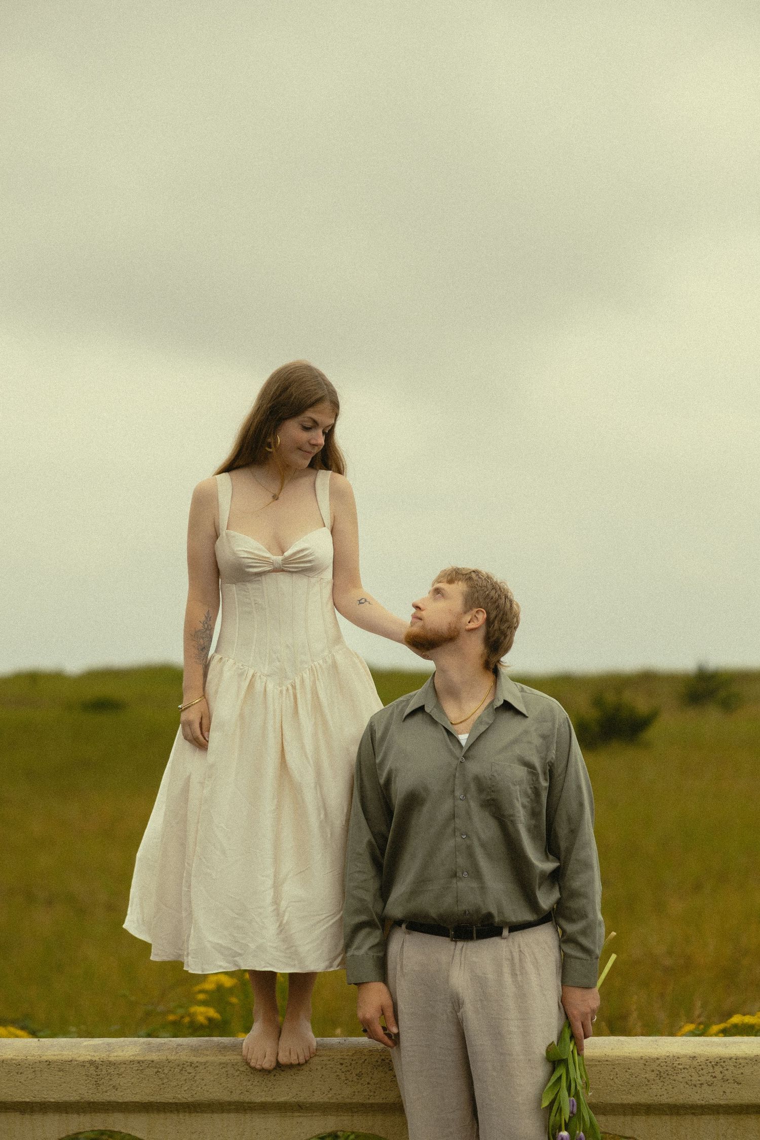 A woman in a white dress stands on a ledge while a man in gray holds flowers in a green field.