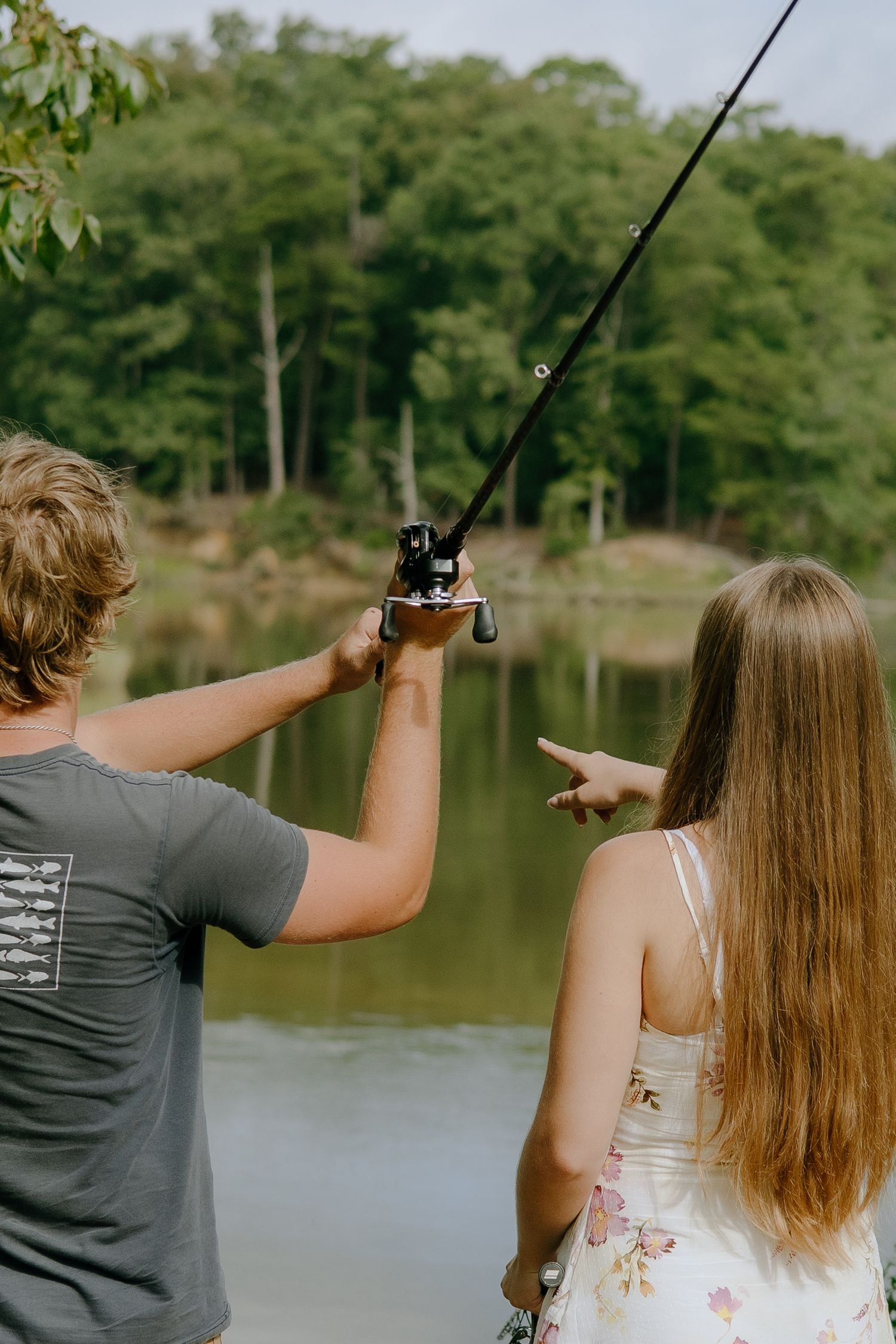 A young couple fishing together by a serene lake surrounded by lush green trees.