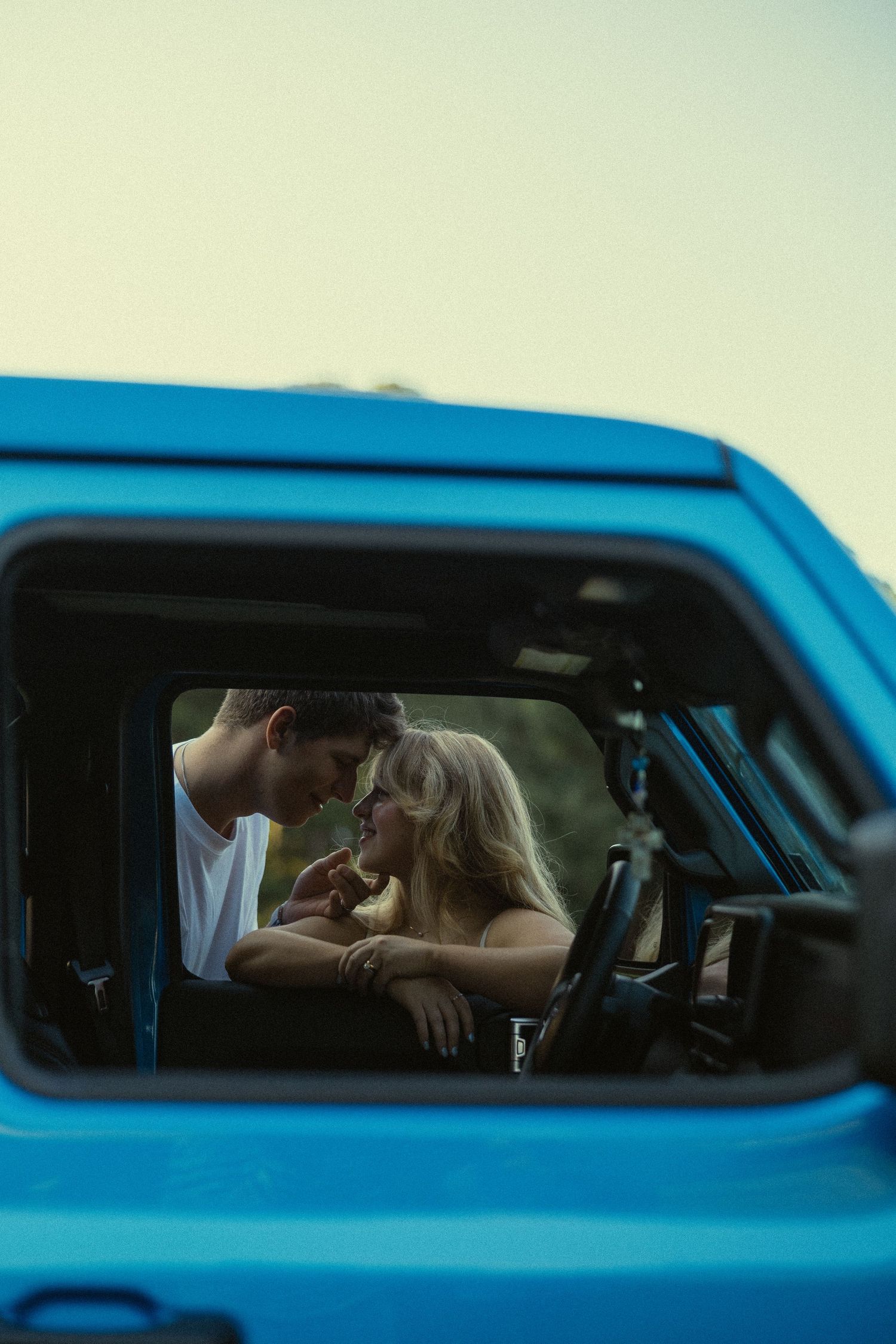 Romantic couple sharing an intimate moment framed through the open window of a blue vintage truck outdoors.