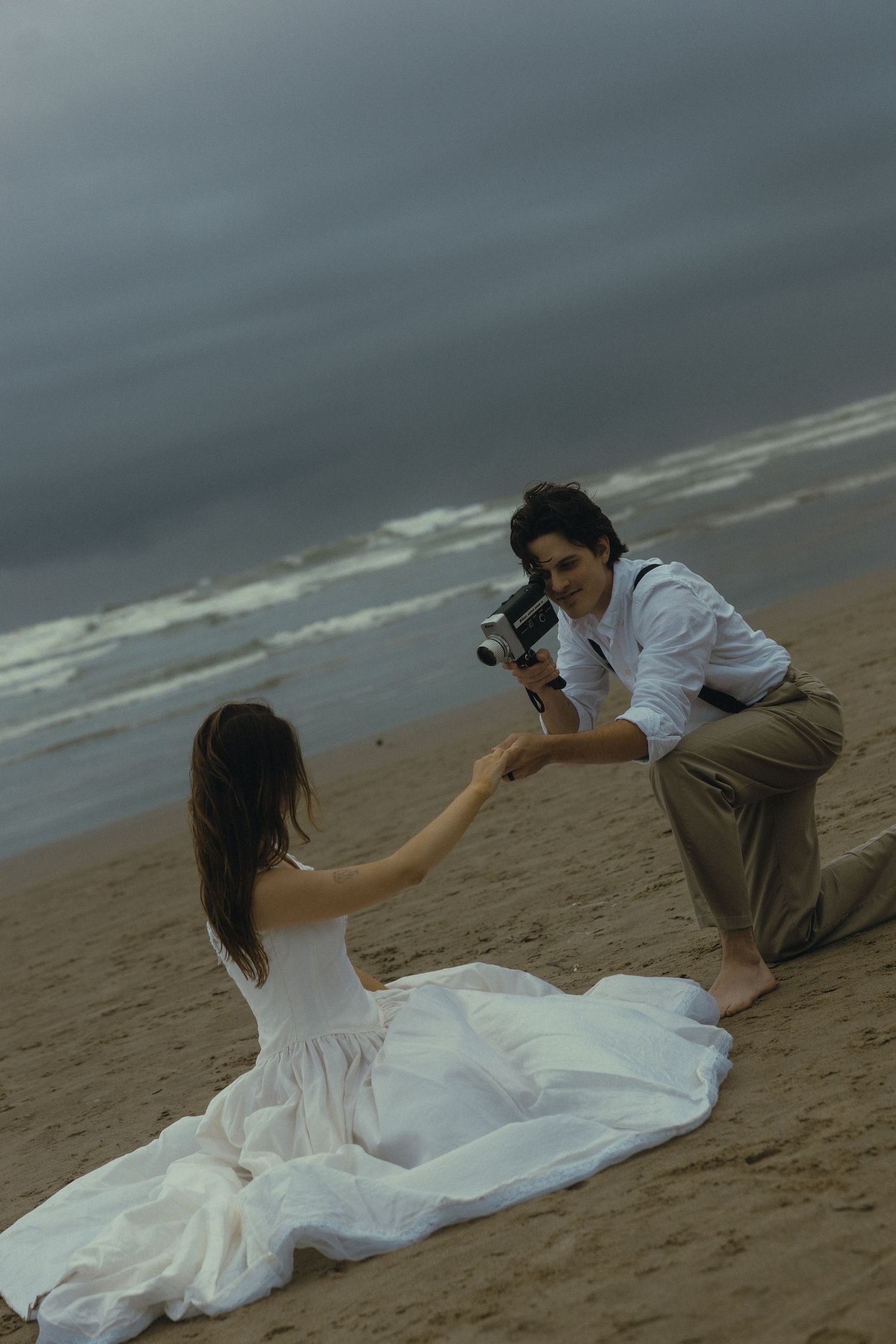 A groom films his bride in a white dress on a sandy beach with stormy skies using a vintage camera.