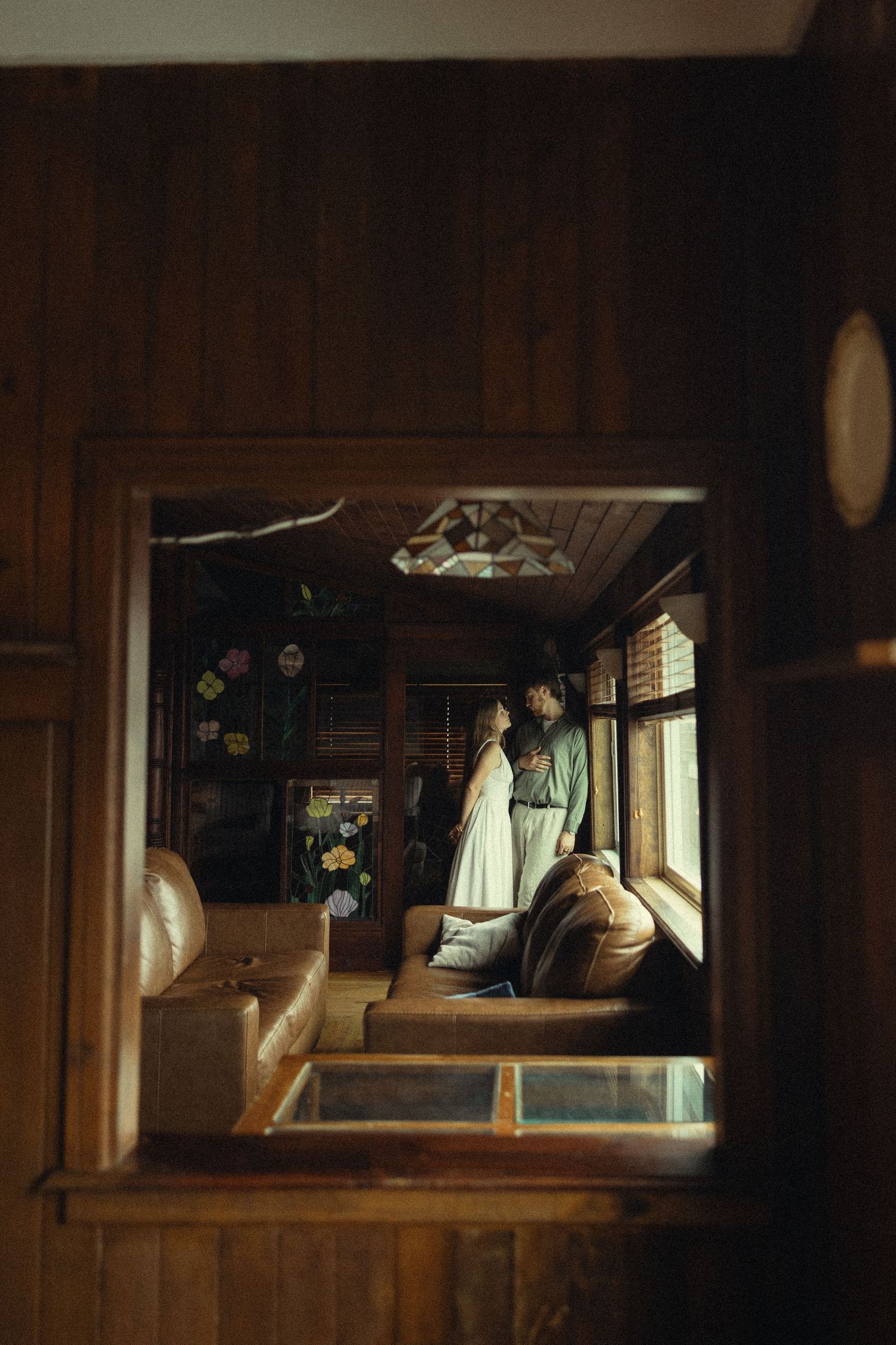 Couple embracing by window in rustic wood-paneled room, viewed through a square opening in warm dim light.