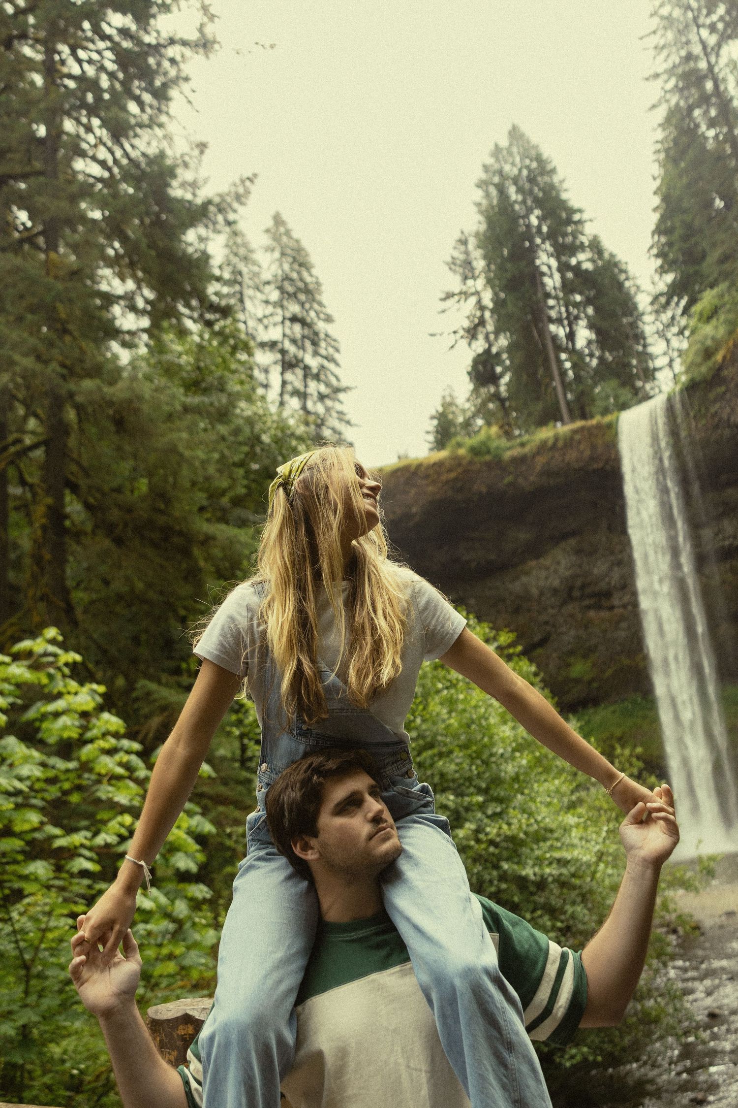 Couple enjoying nature, woman sitting on man's shoulders near a waterfall surrounded by lush green forest.