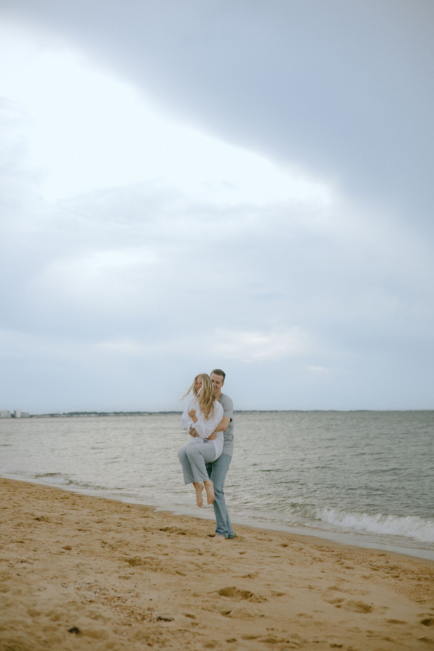 A romantic moment shared on a sandy beach with cloudy skies and ocean waves in the background.