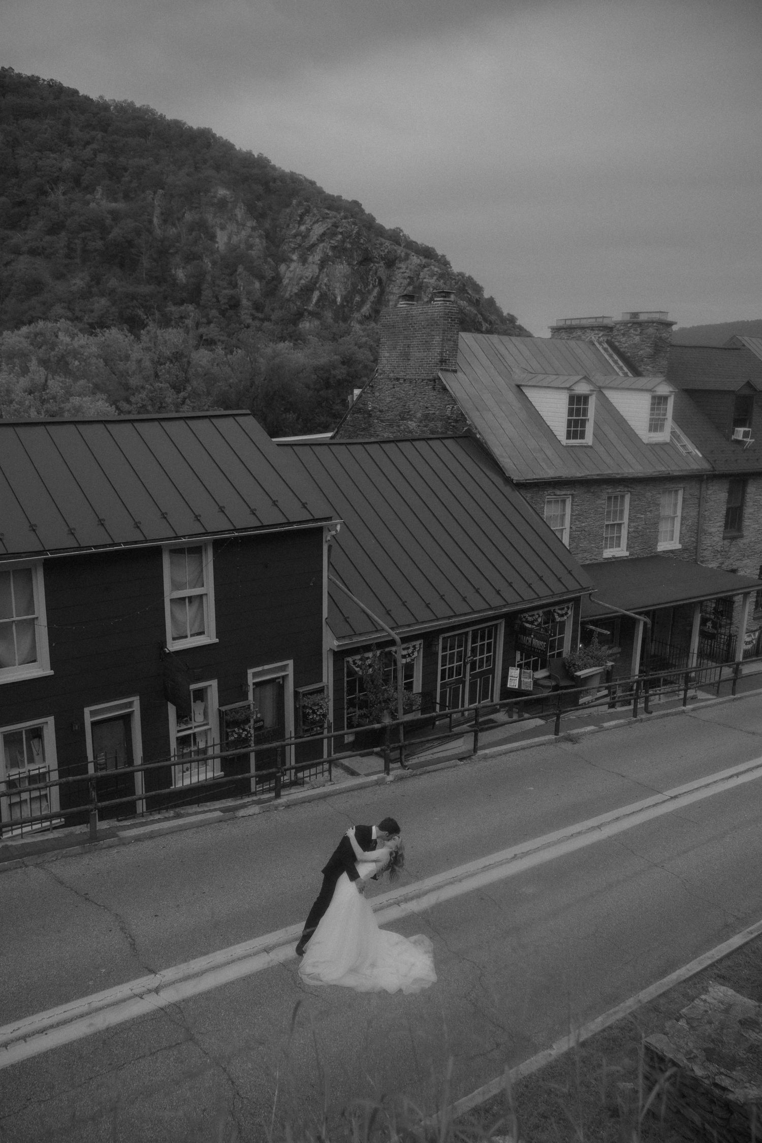 Black and white photo of a couple sharing a romantic dip kiss on a small town street with historic buildings and a mountain backdrop.