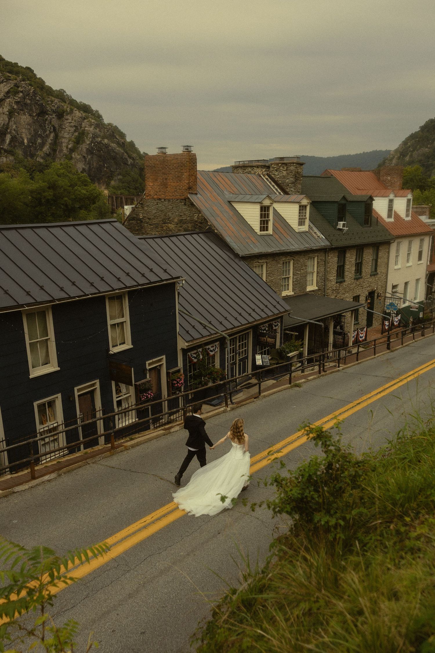 Bride and groom cross a historic small-town street in Harpers Ferry, WV, with mountain backdrop on their wedding day.