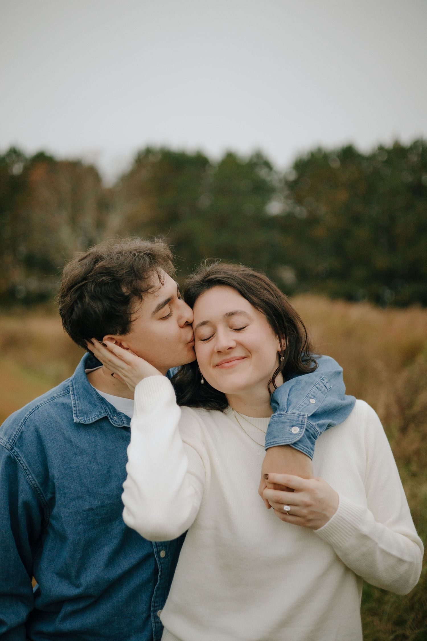 A couple shares a tender kiss outdoors in an autumn field, the woman smiling warmly while holding a coffee cup.