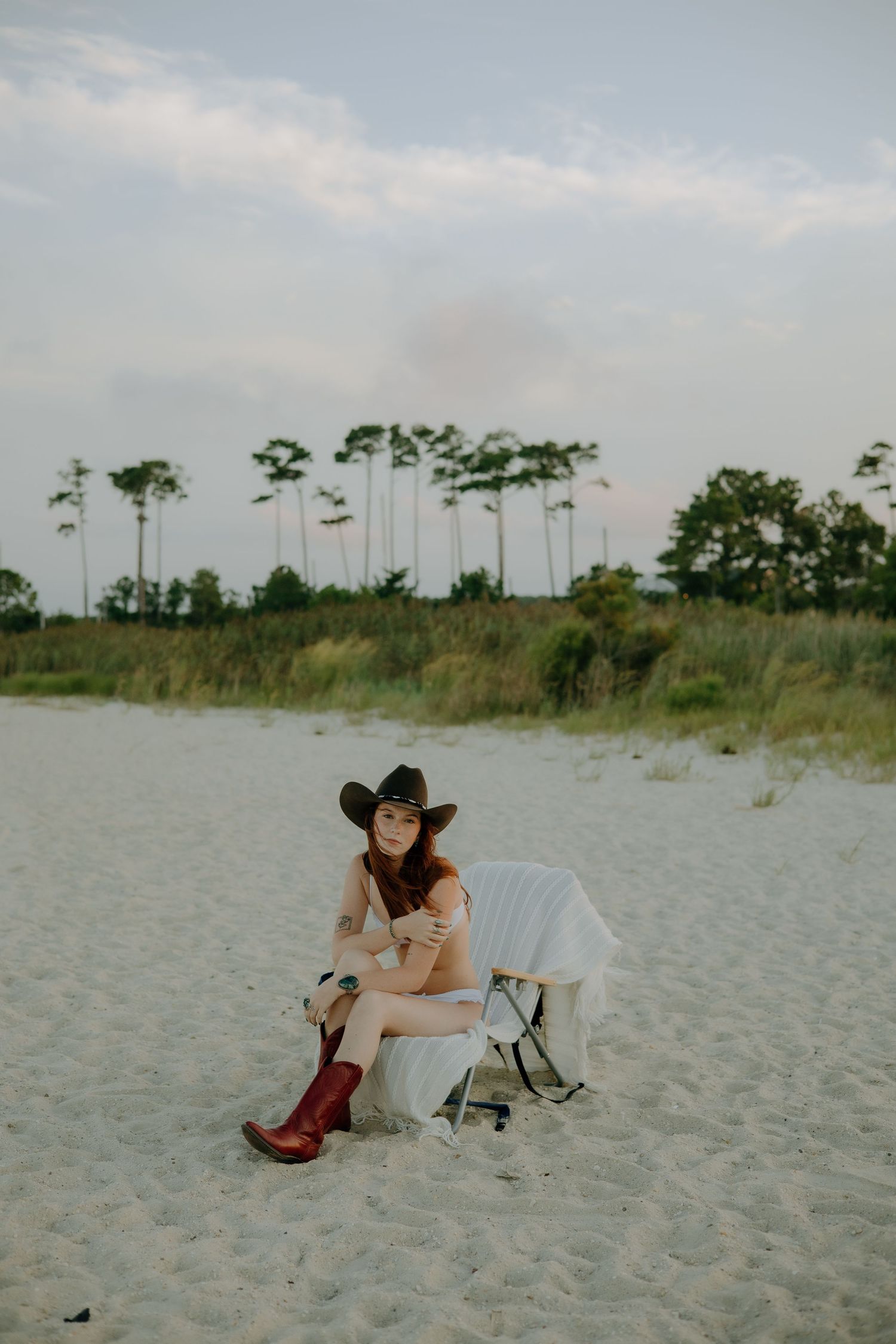 Woman in cowboy hat and red boots sits on sandy beach with white dress, trees in background.