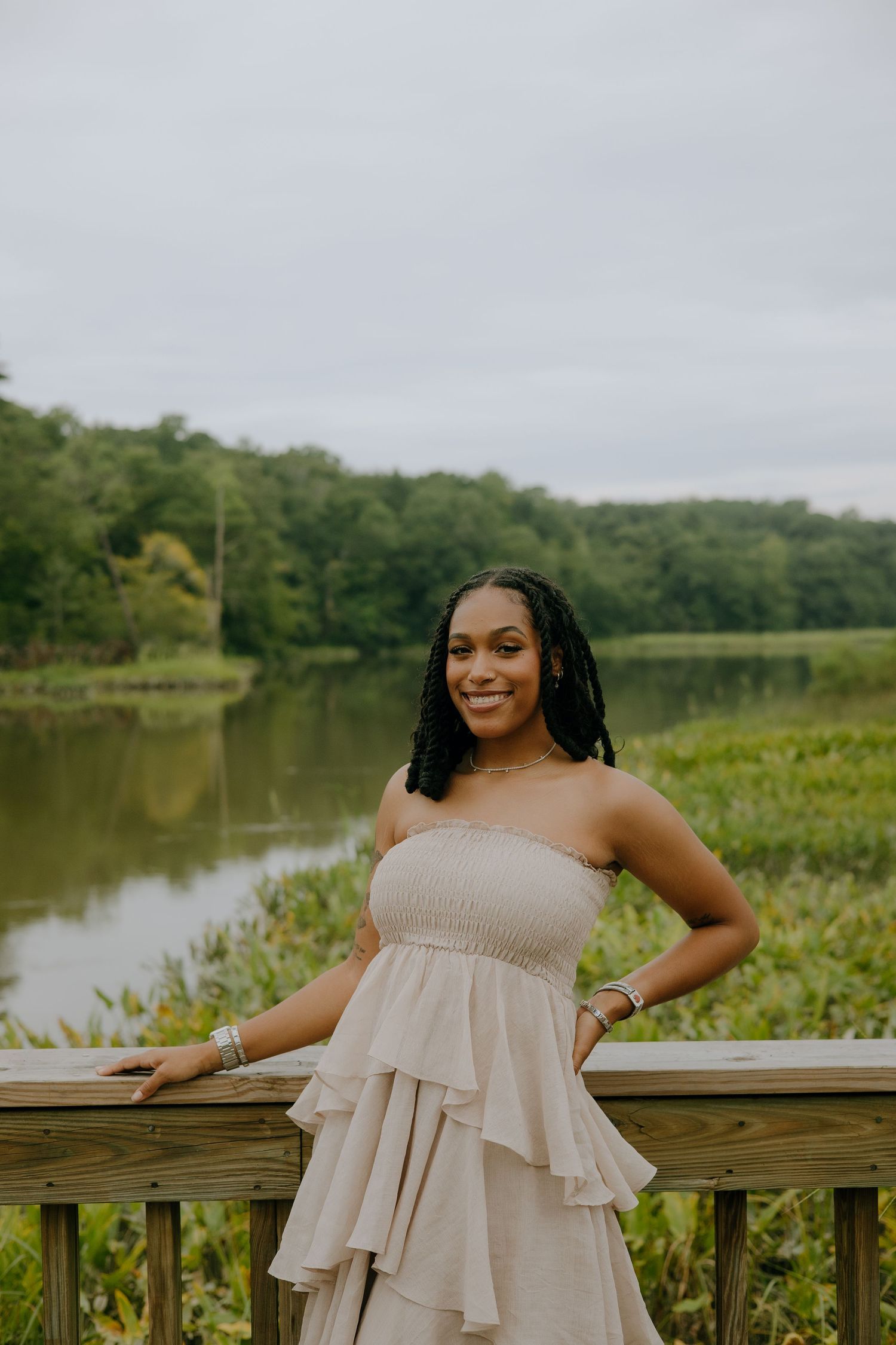 Young woman in a beige ruffled strapless dress posing on a wooden deck overlooking a serene lake and lush greenery.