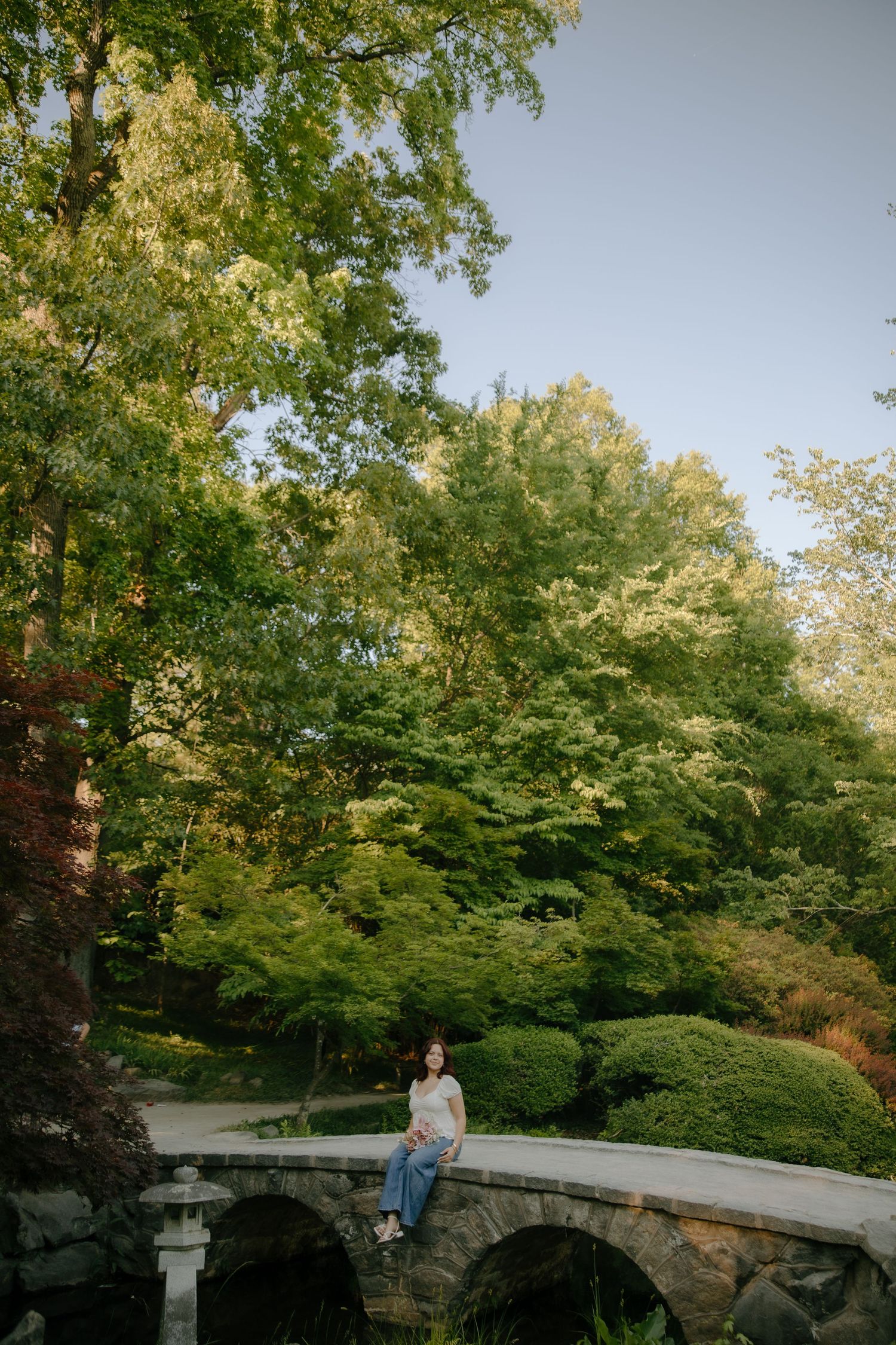 Woman sitting on a stone arch bridge surrounded by lush green trees in a serene garden park setting.