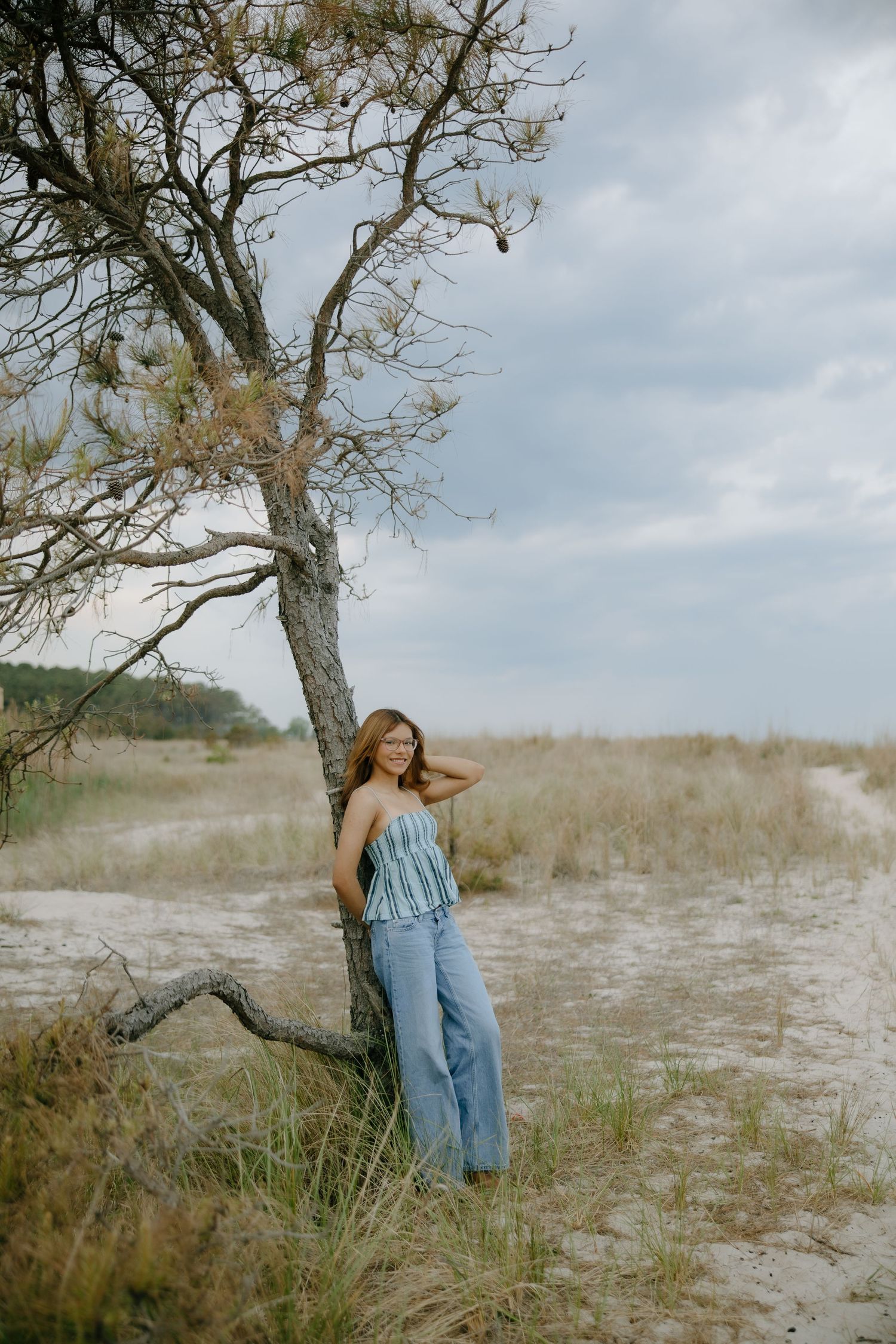 Young woman in blue jeans and striped top leaning against a bare tree on a sandy beach under cloudy skies.