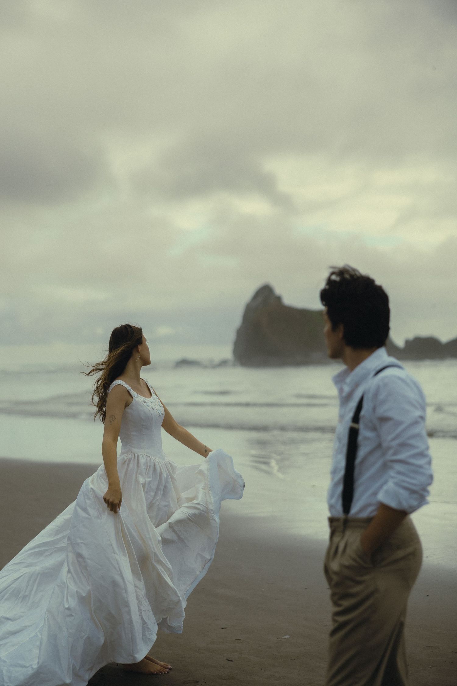 A bride in a flowing white dress stands on a windy beach as her groom watches her admiringly.
