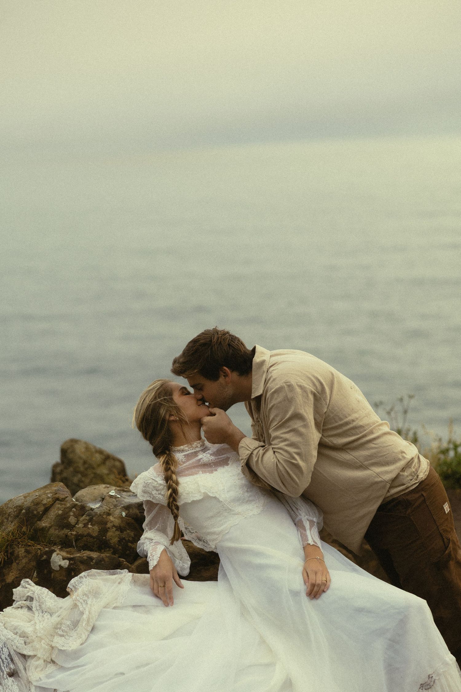 Romantic couple sharing a kiss on seaside rocks, bride in white lace gown with groom in beige shirt.