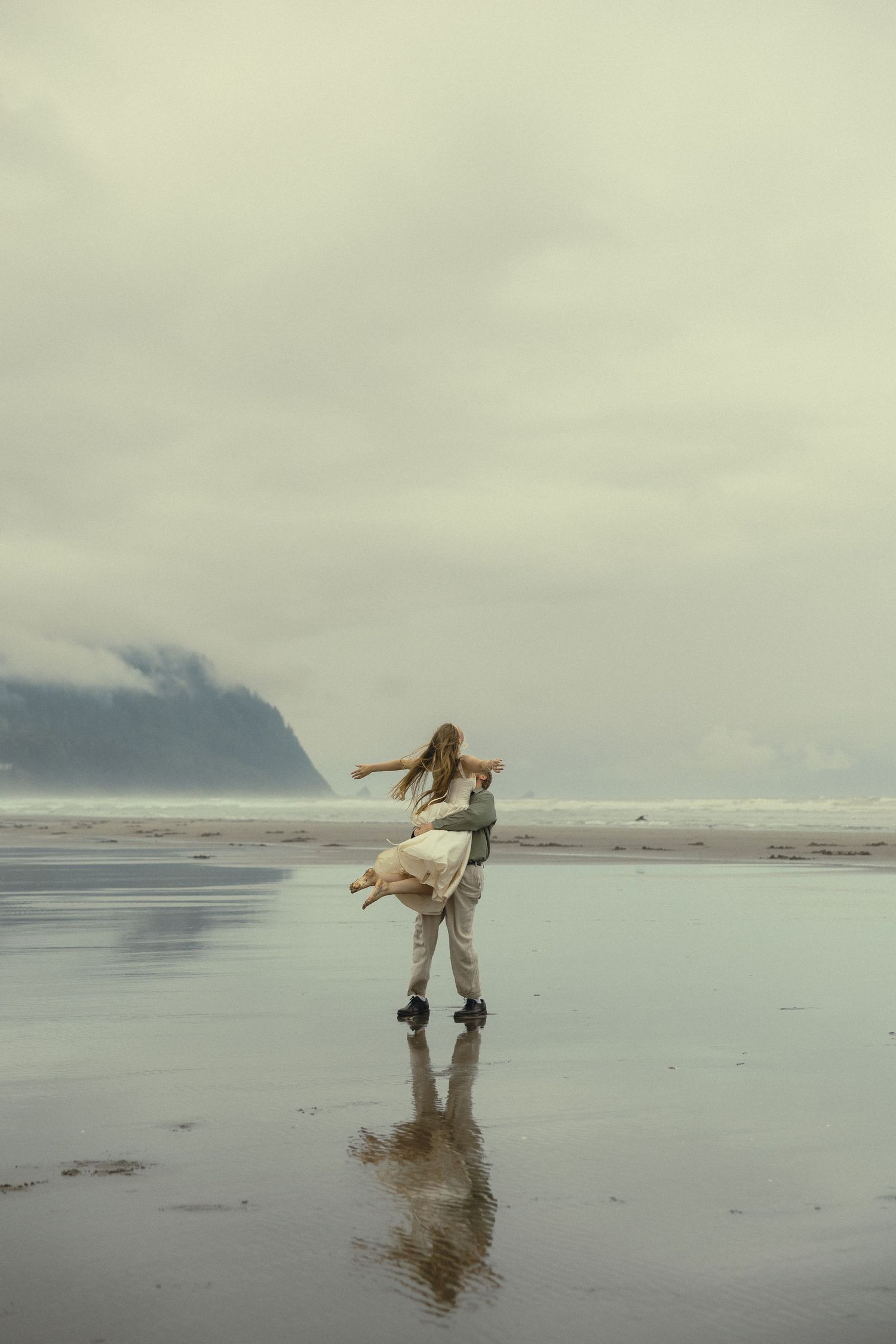 A couple dances on a misty beach with mountain reflections on wet sand.