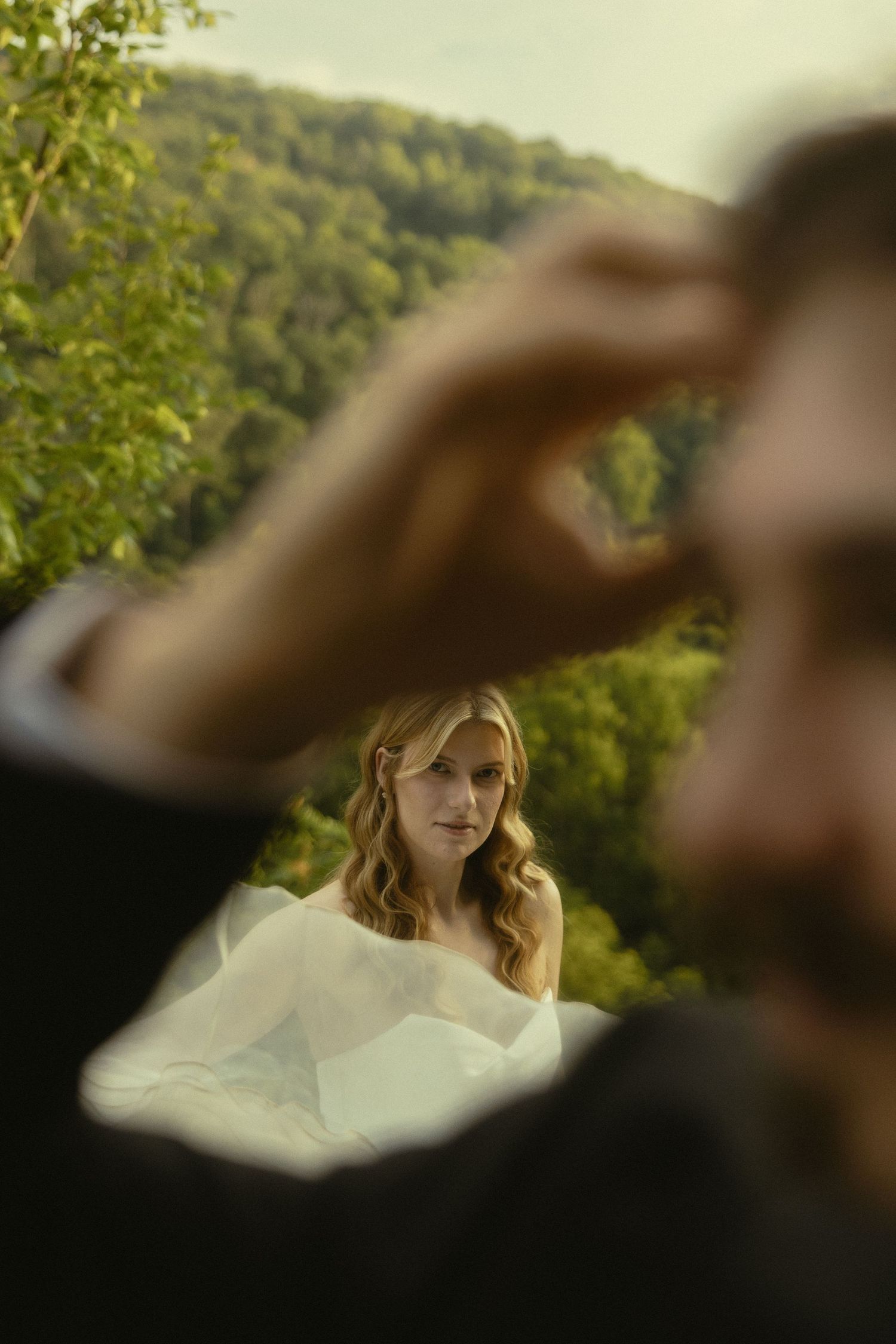 Bride in white gown stands before lush green hillside, viewed through groom's blurred foreground figure.